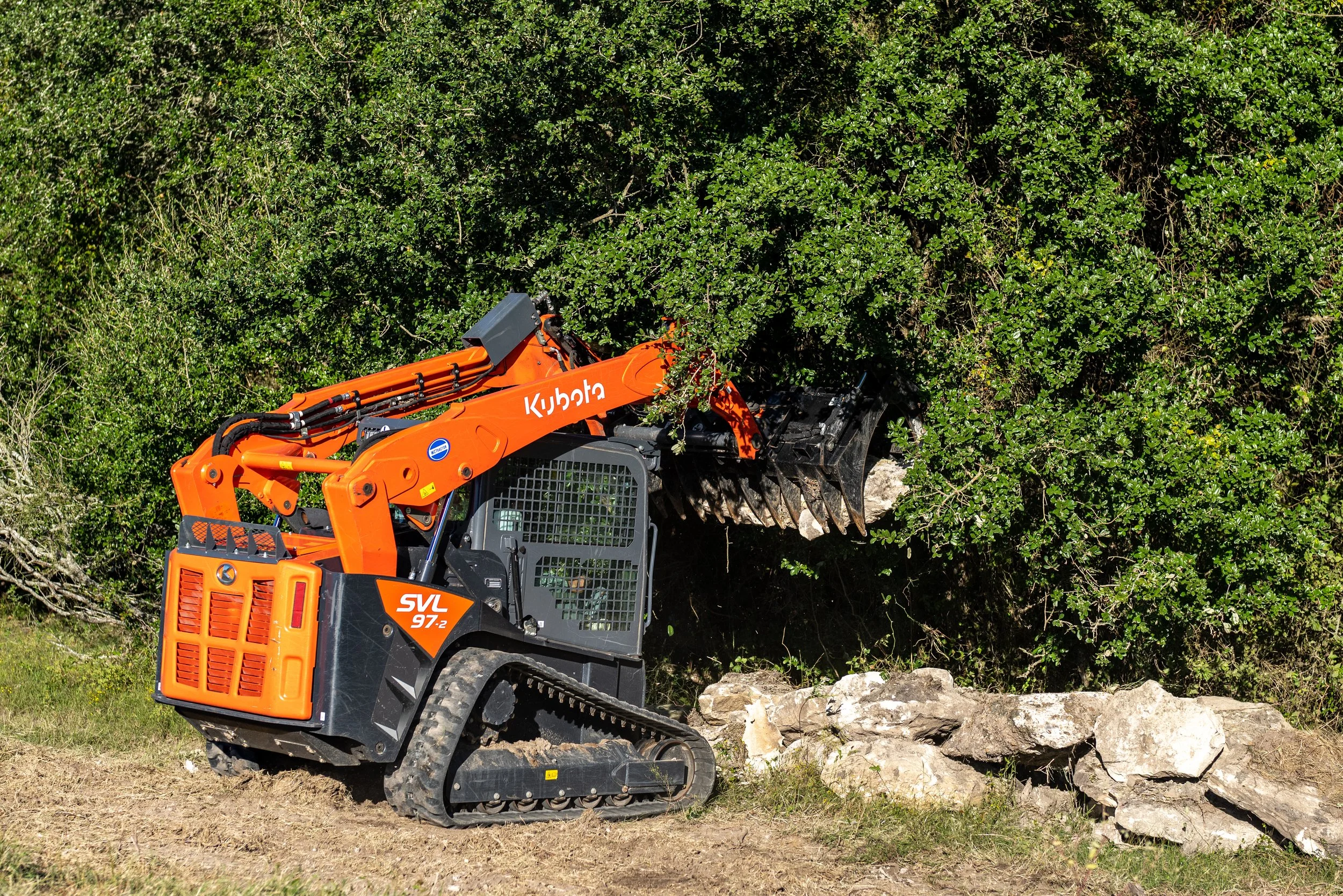 Orange excavator with front bucket crane moving rocks near dense green foliage.