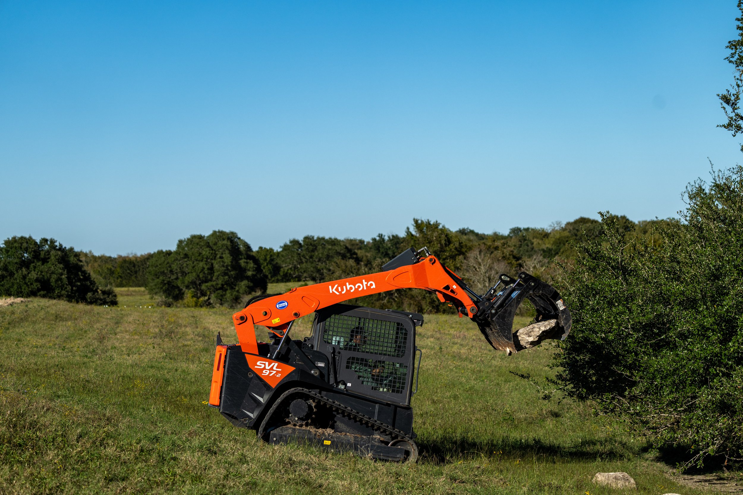 Orange Kubota skid steer loader on grass, lifting a rock with a grapple attachment, against a backdrop of trees and blue sky.