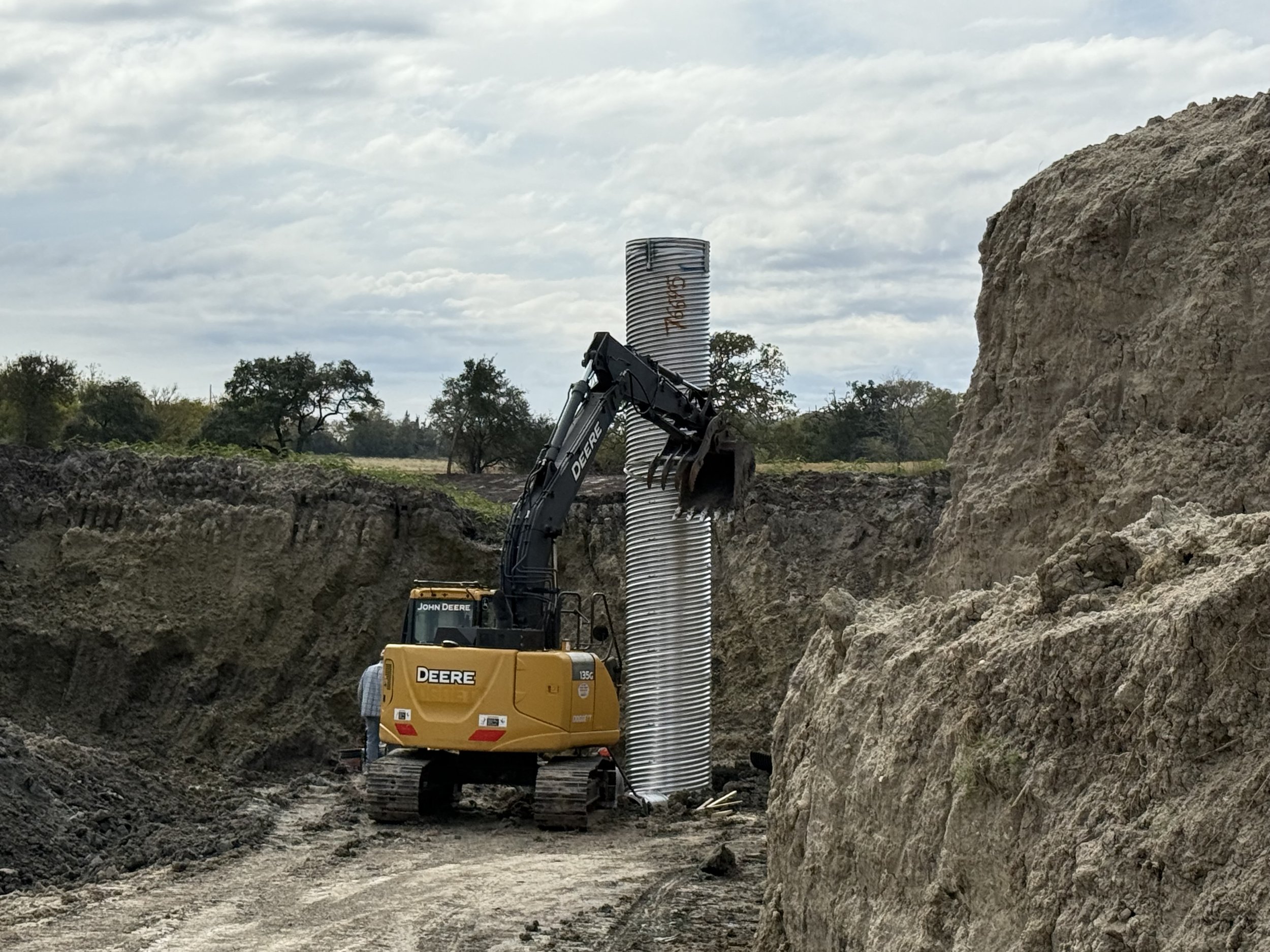 Construction excavator moving a large corrugated metal pipe in a dirt excavation site with rocky walls and trees in the background, provided by G-Quip Services LLC in the Brazos Valley, Texas.