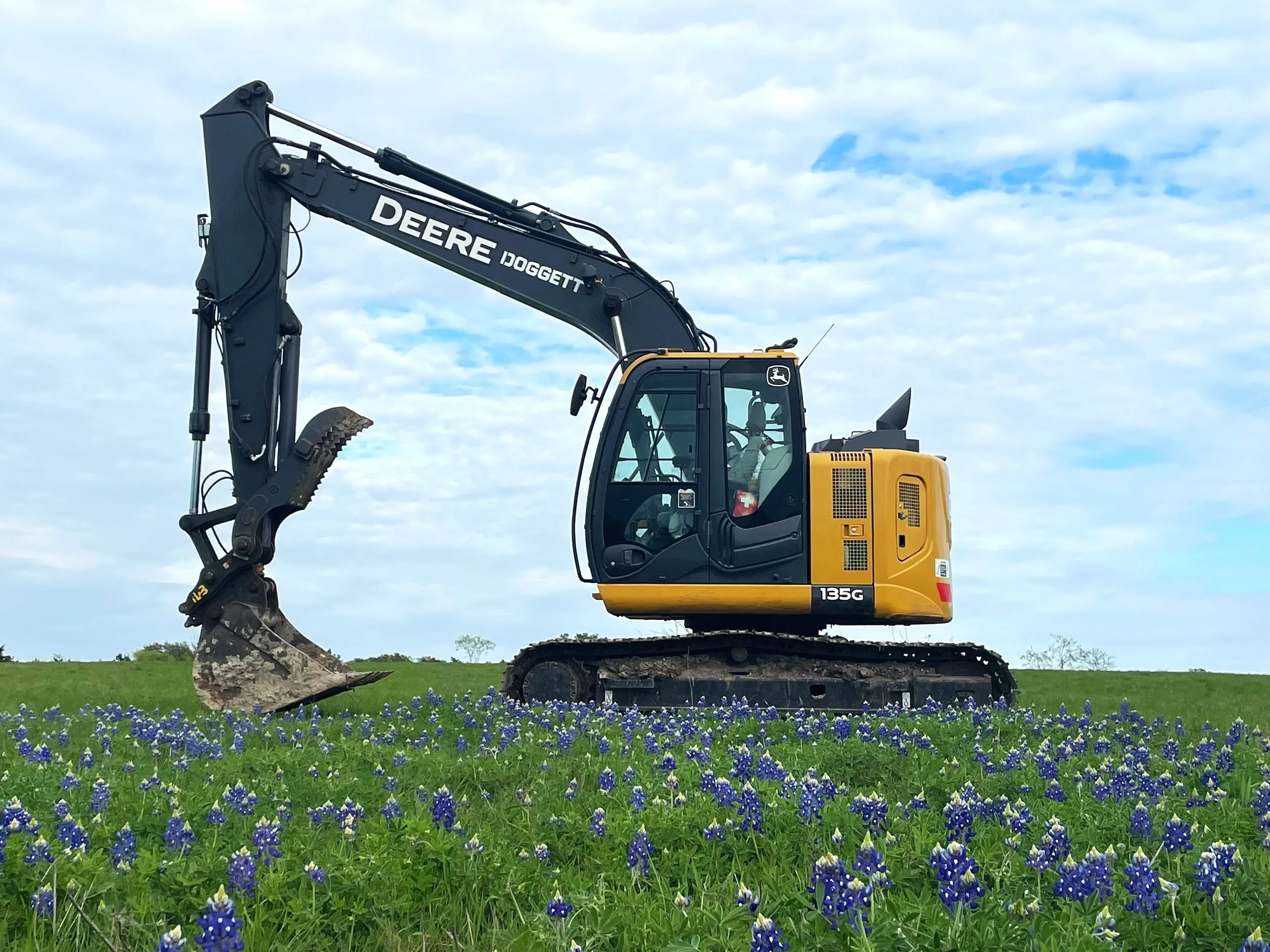 Yellow and black John Deere excavator working in a field of bluebonnet flowers under a partly cloudy sky, in the Brazos Valley, Texas.
