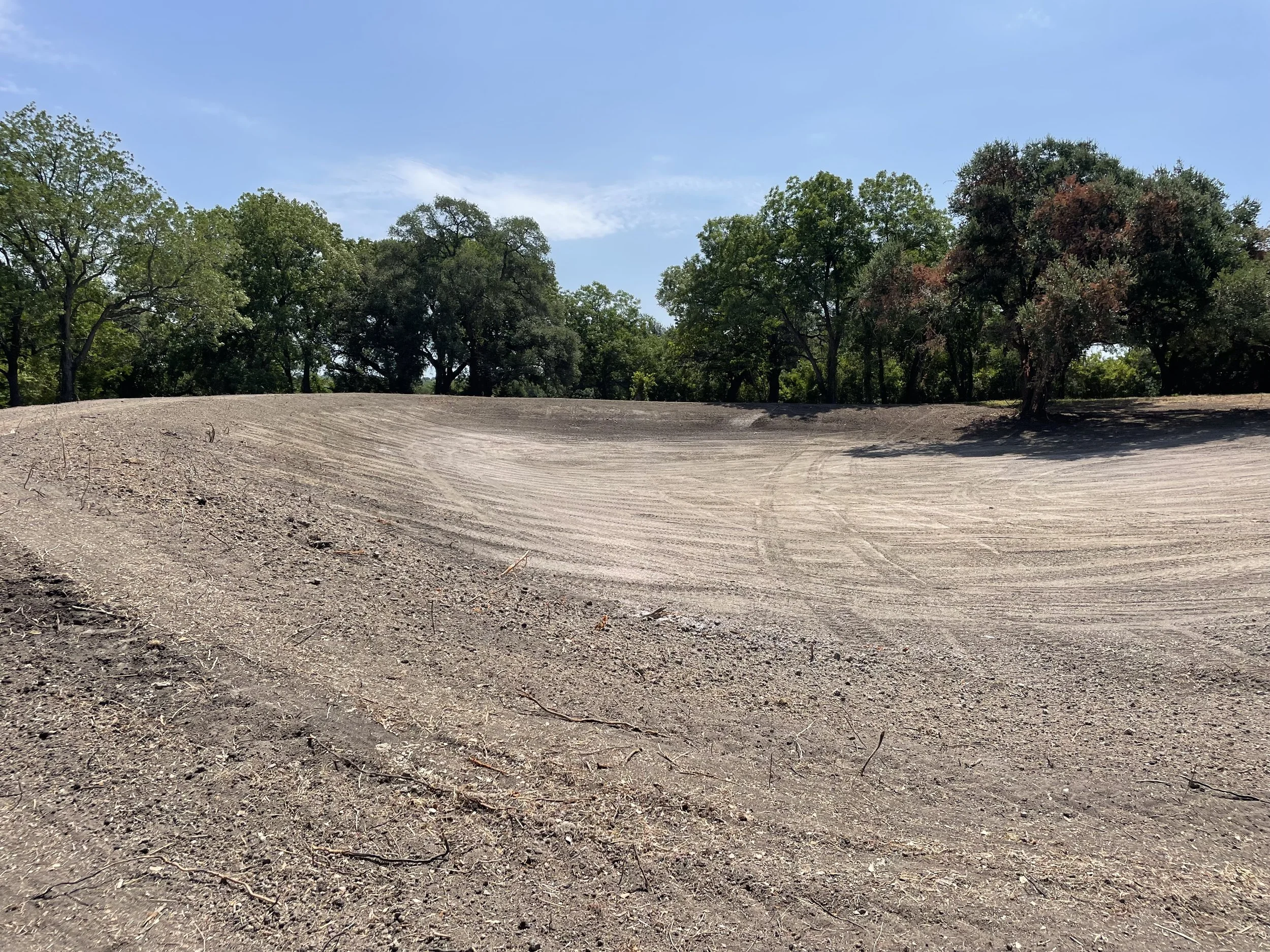 A landscaped lake and pond with a cleared, dirt area surrounded by green trees under a blue sky, provided by G-Quip Services LLC in the Brazos Valley, Texas.