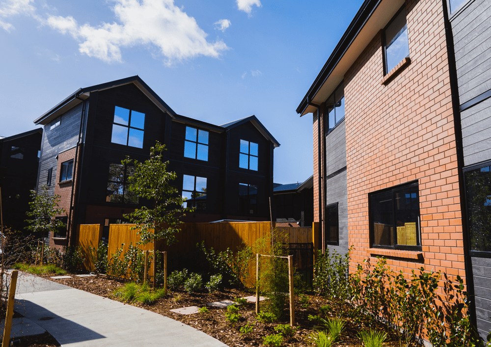 49 Te Kanawa Crescent: A modern residential neighborhood with contemporary multi-story townhouses, some with dark siding and others with brick exteriors, under a blue sky with some clouds. There are small trees and a landscaped yard in the foreground.