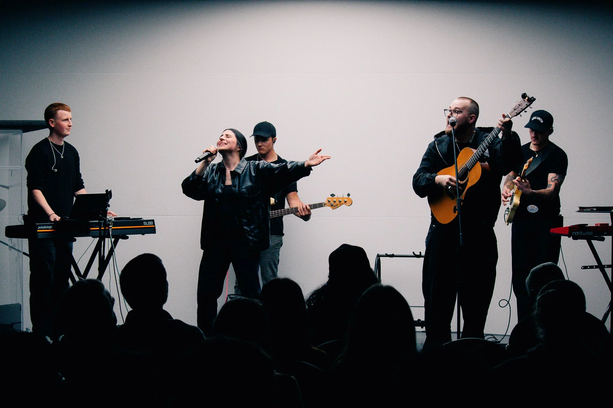 A group of five musicians performing on stage with various instruments, including a keyboard, bass guitar, acoustic guitar, and electric guitar, with a woman singing into a microphone in front of an audience.