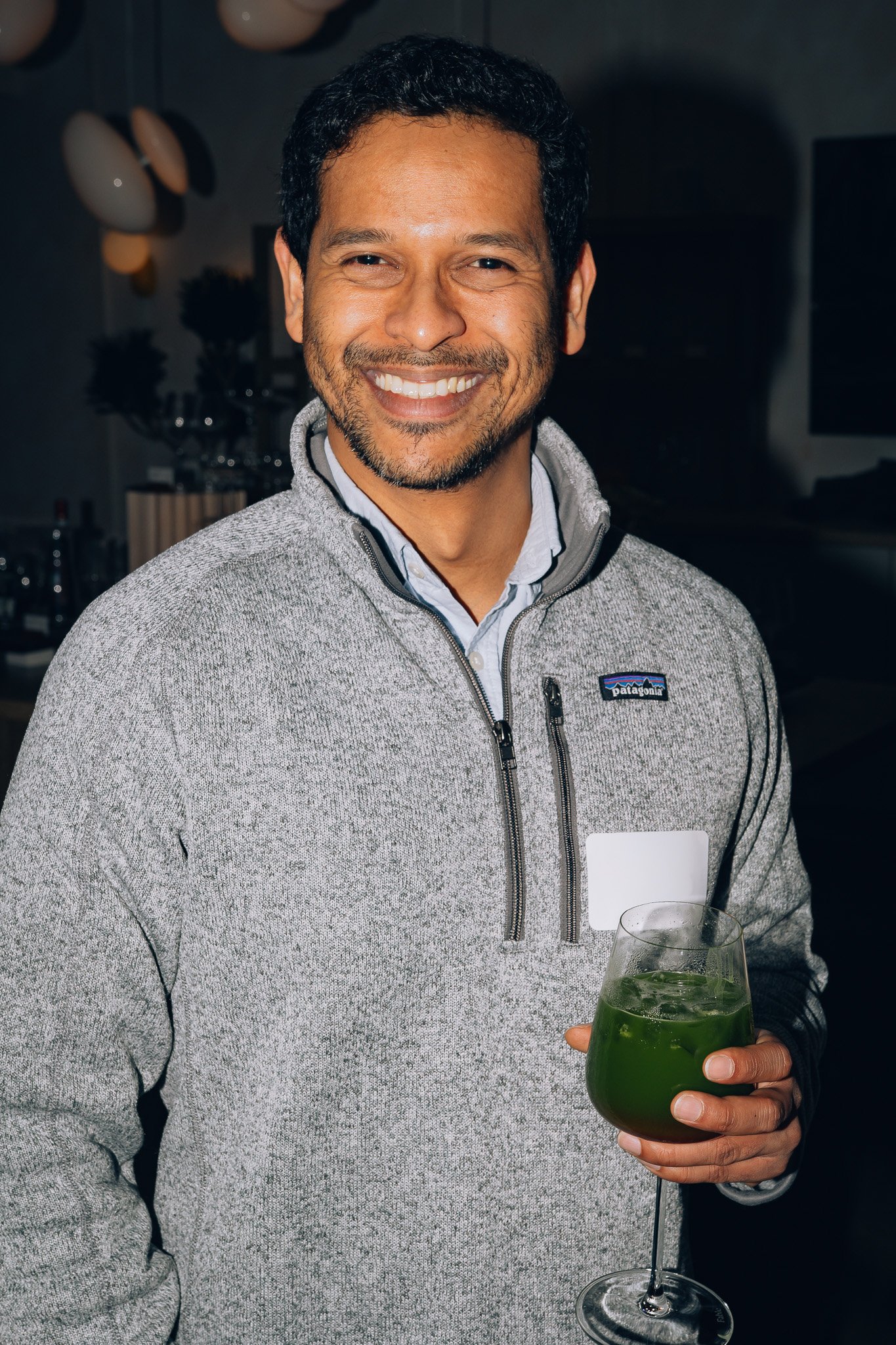 A man smiling while holding a green drink in a stemmed glass with lime slices, in a dimly lit indoor setting.