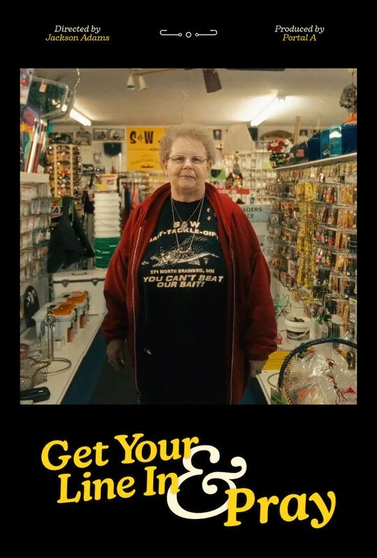 An elderly woman standing inside a store, wearing glasses, a black T-shirt with fishing-related text, a red jacket, and a necklace. The store shelves behind her are stocked with various items.