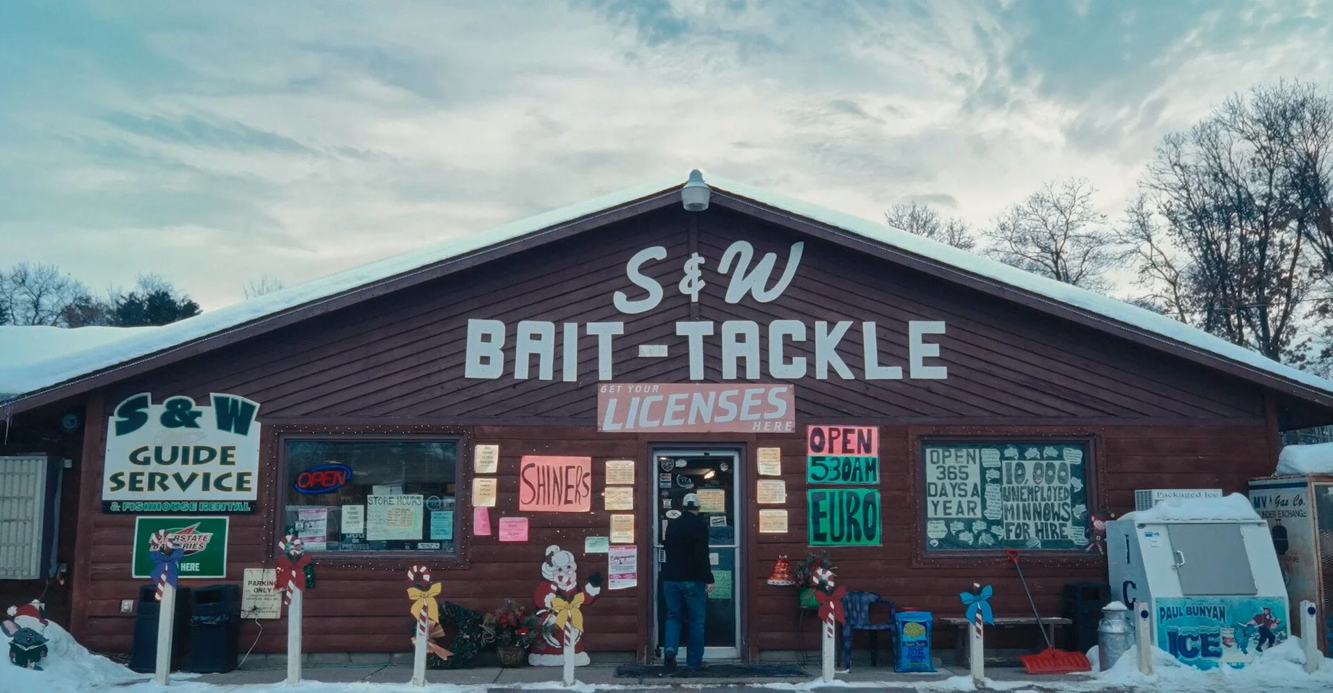 Front of a wooden bait shop with signs advertising licenses, hours, and snow outside, decorated with Christmas-themed decorations.