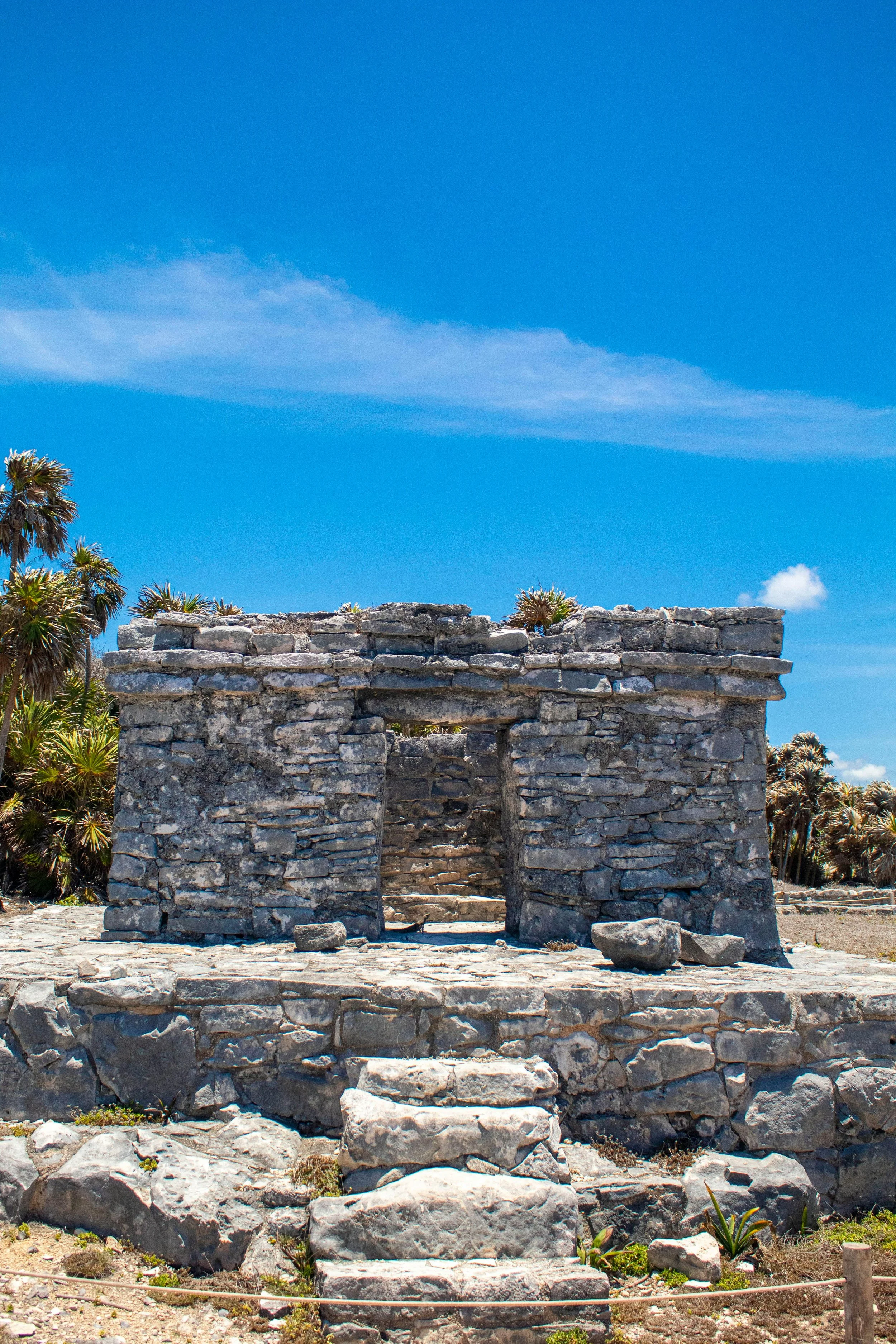 Ancient stone structure with steps leading up to a doorway, surrounded by palm trees, under a bright blue sky.