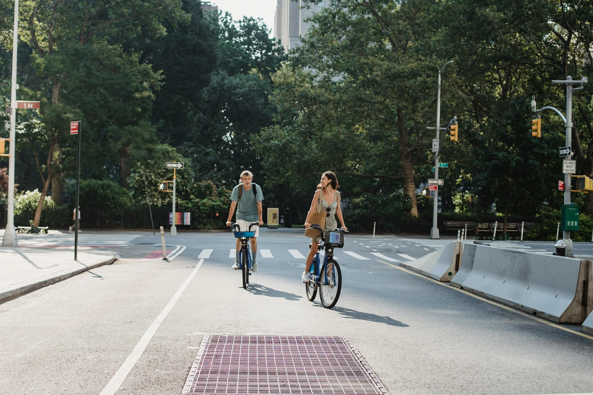 Two women riding bicycles on a sunny city street with trees, traffic lights, and crosswalks.