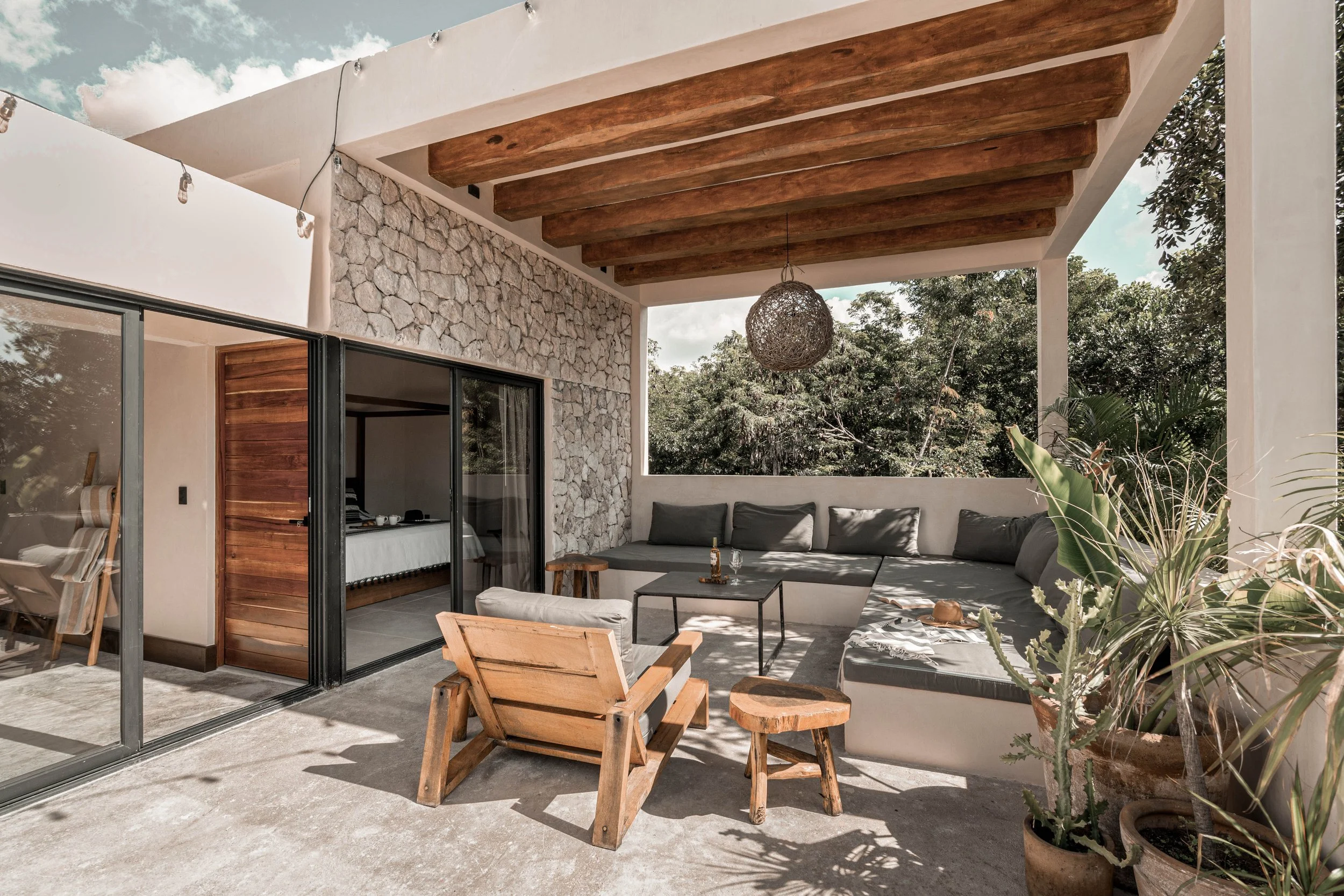 Outdoor patio with a large gray sectional sofa, a wooden armchair, a small wooden stool, and a black metal coffee table. Potted plants are on the right side, and a stone wall is behind the sofa. A woven hanging light fixture is suspended from wooden ceiling beams, with greenery and trees visible in the background.
