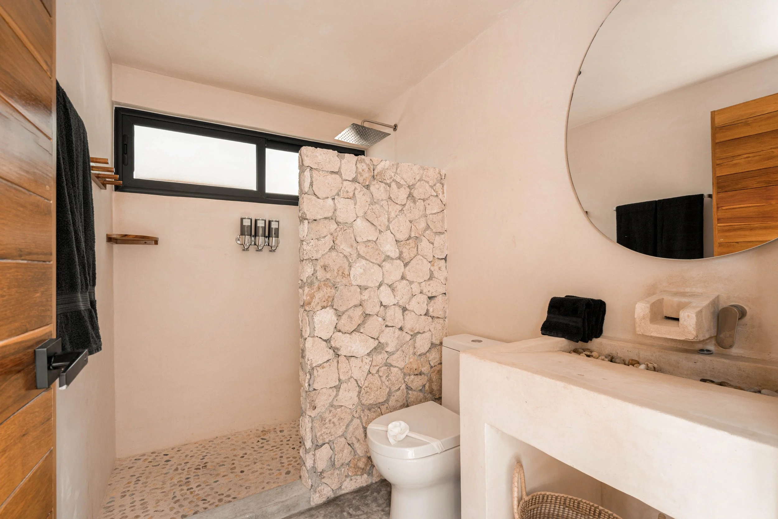 Bathroom with a stone partition, a toilet, a circular mirror, a stone sink, a black towel, and a shower area separated by the stone wall.