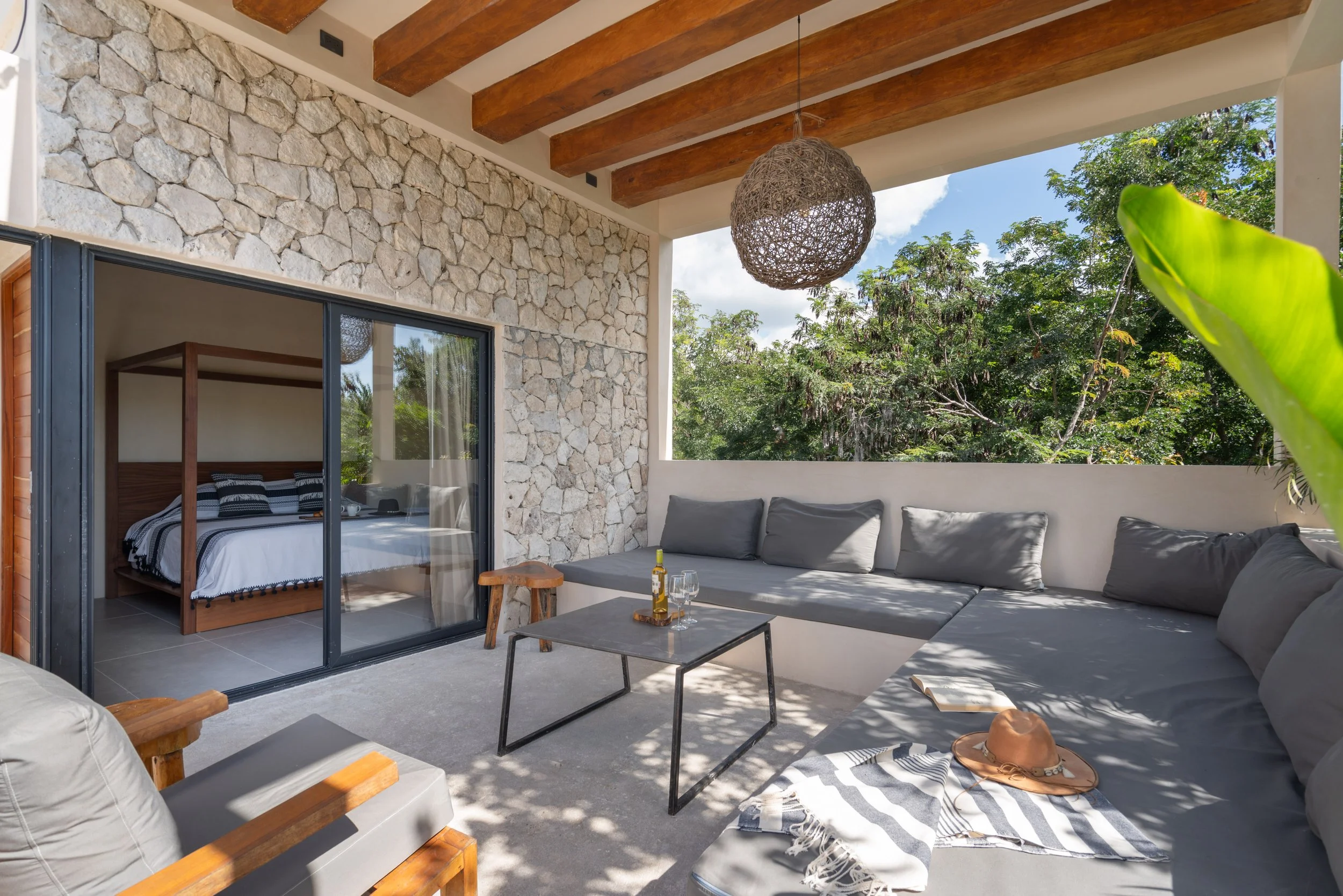 Outdoor patio with built-in gray cushioned seating, a black metal coffee table with a bottle and glass, wooden accents, stone wall, large trees in the background, and a bedroom with a wooden bed and black and white bedding visible through sliding gla