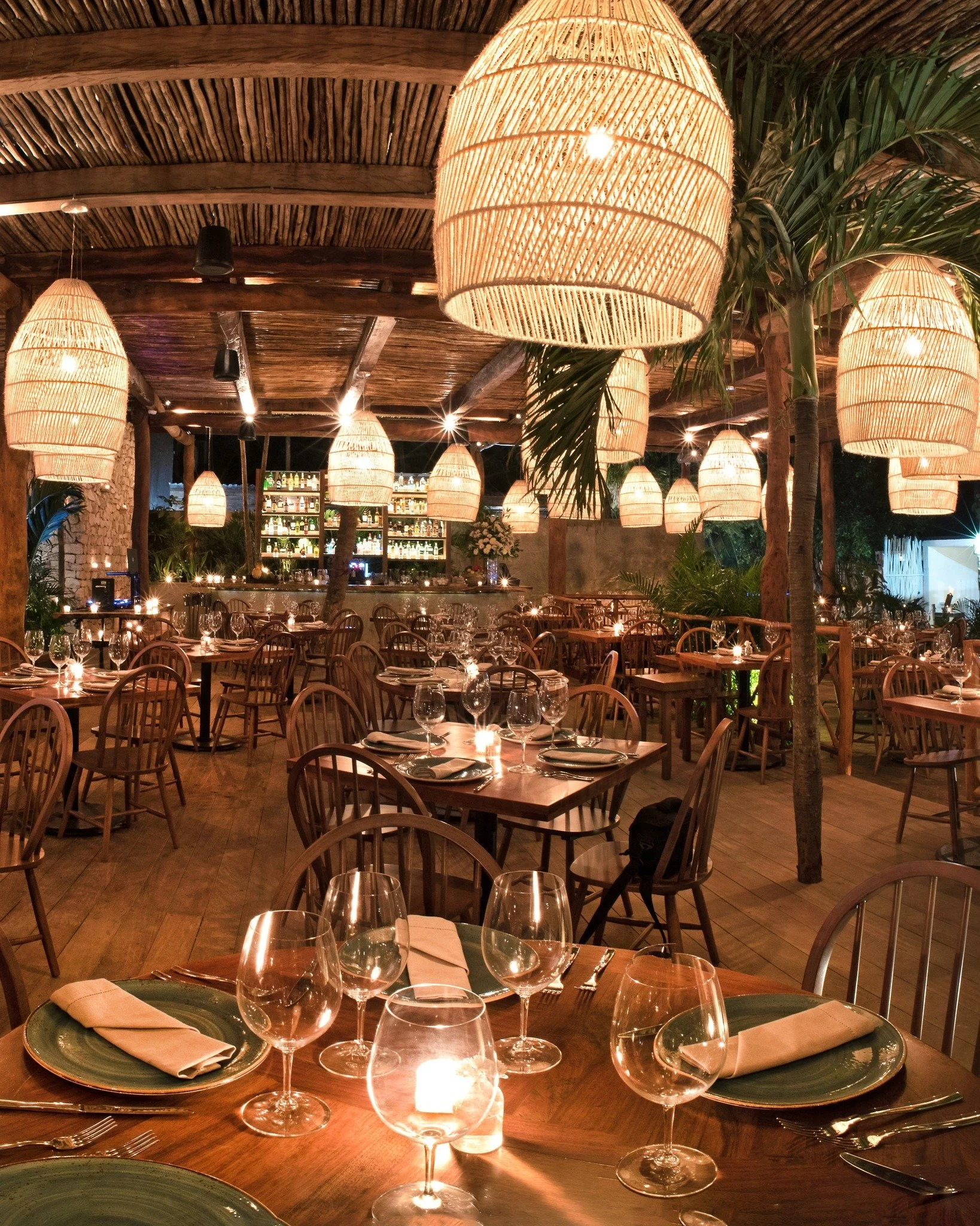 Elegant restaurant with wooden tables set for dining, wine glasses, candles, and warm hanging lanterns.