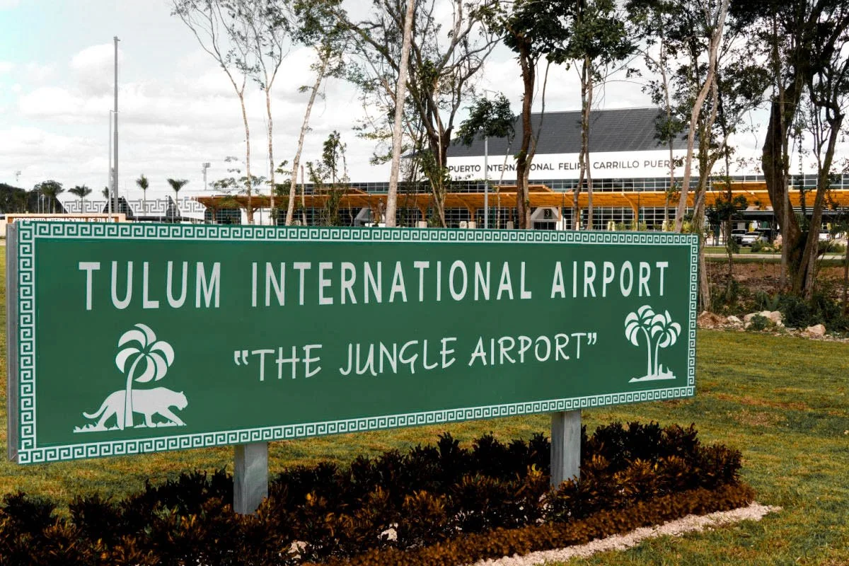 Sign for Tulum International Airport, also known as The Jungle Airport, featuring a lion and palm tree graphic, with airport building and trees visible in the background.