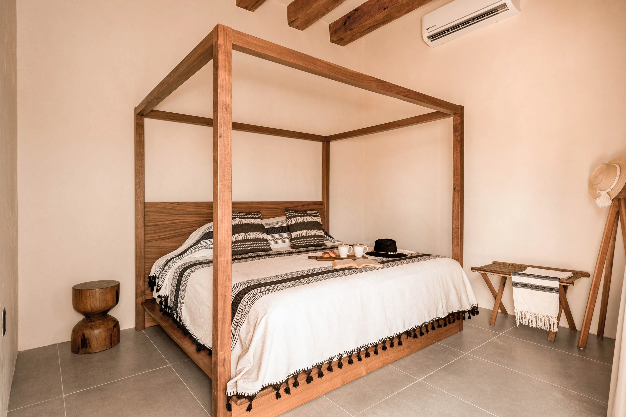 A bedroom with a wooden four-poster bed, white and black patterned bedding, and pillows. There is a small wooden side table on the left, and on the right, a bench with a woven seat, a towel, and a straw hat hanging on a stand. An air conditioner is mounted on the wall above.
