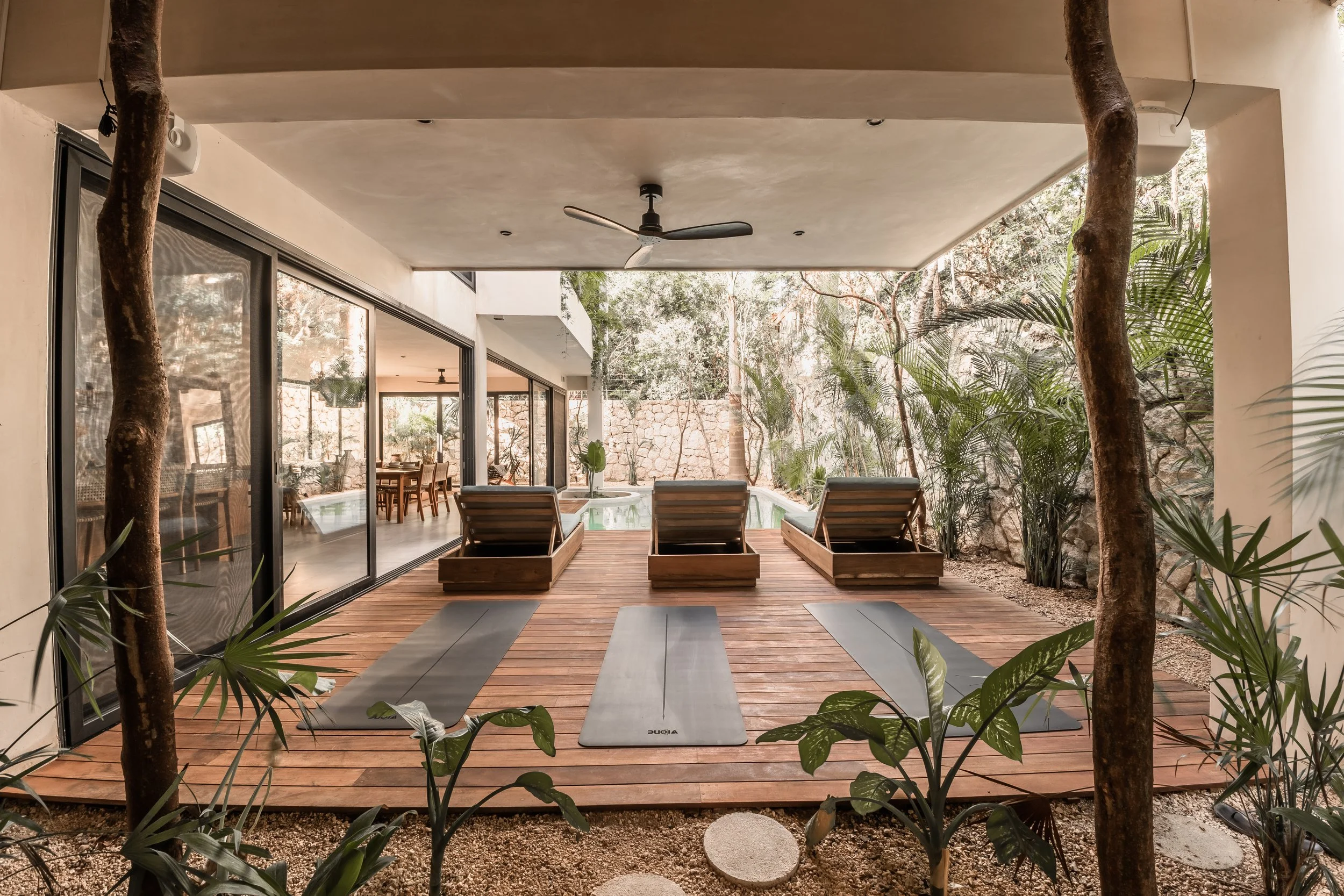 Indoor-outdoor space with yoga mats, lounge chairs, plants, and glass sliding doors leading to a dining area inside, with a pool and trees outside.