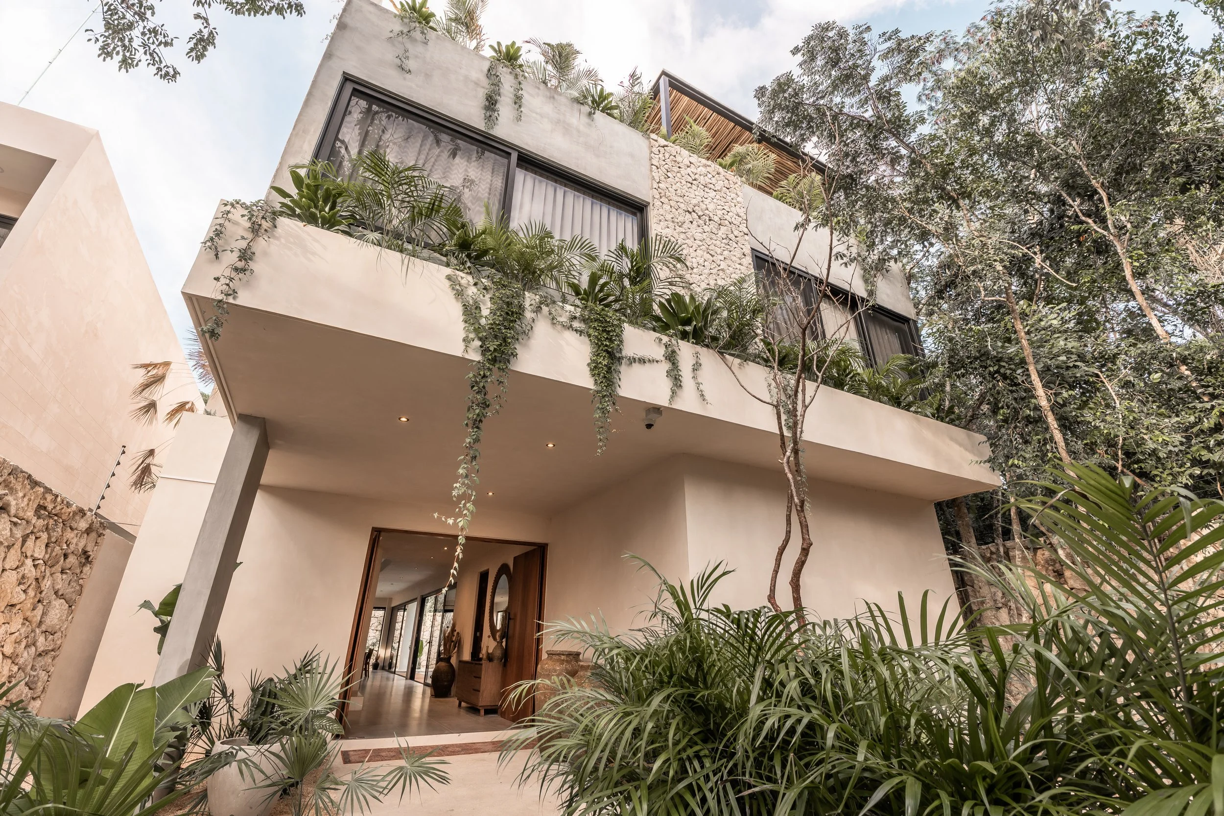 Modern house with a large balcony, lush greenery, and large windows, surrounded by trees.