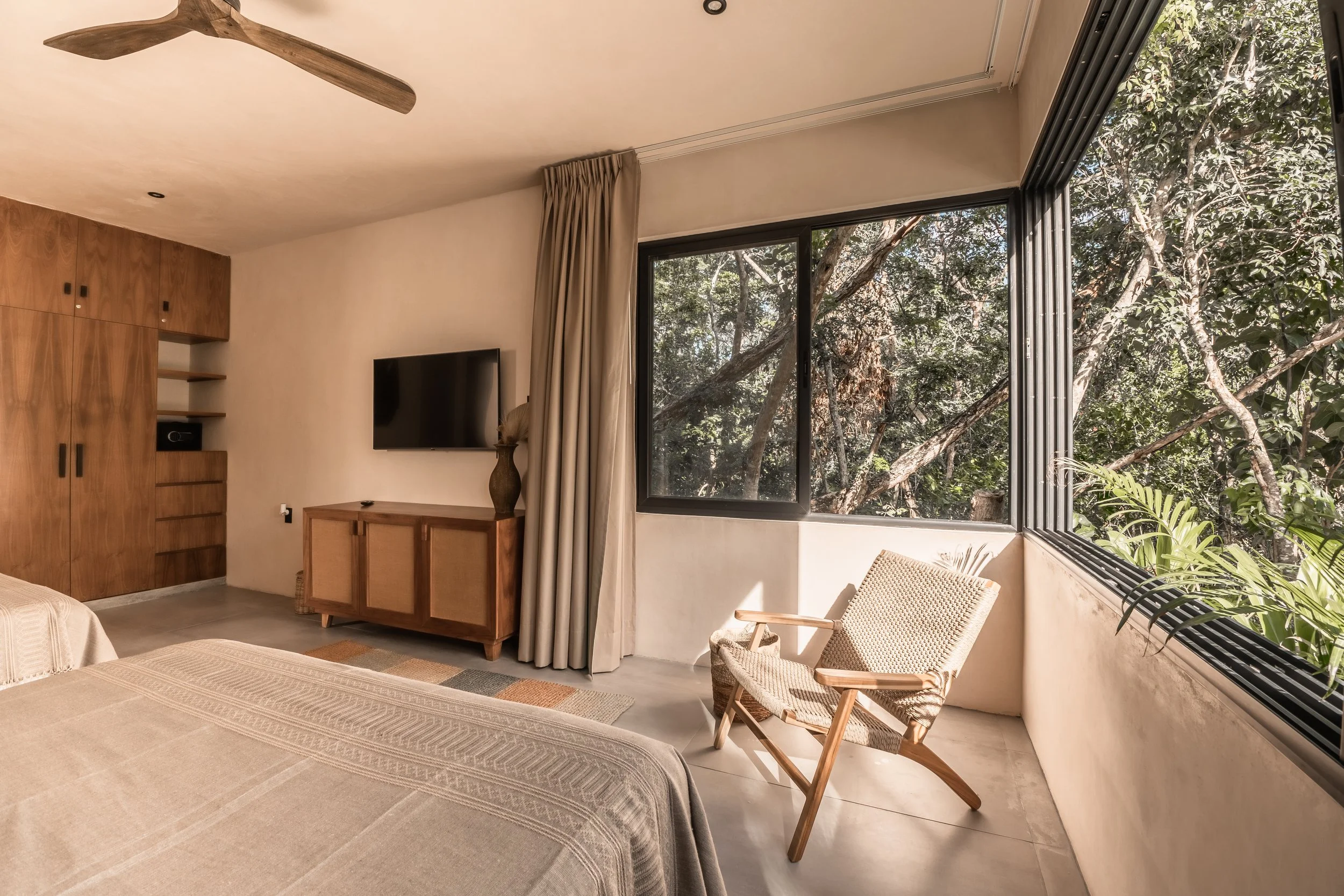 A bedroom with beige bedsheets, a flat-screen TV on a wooden cabinet, a wooden wardrobe, a beige armchair, and large windows showing a view of trees outside.