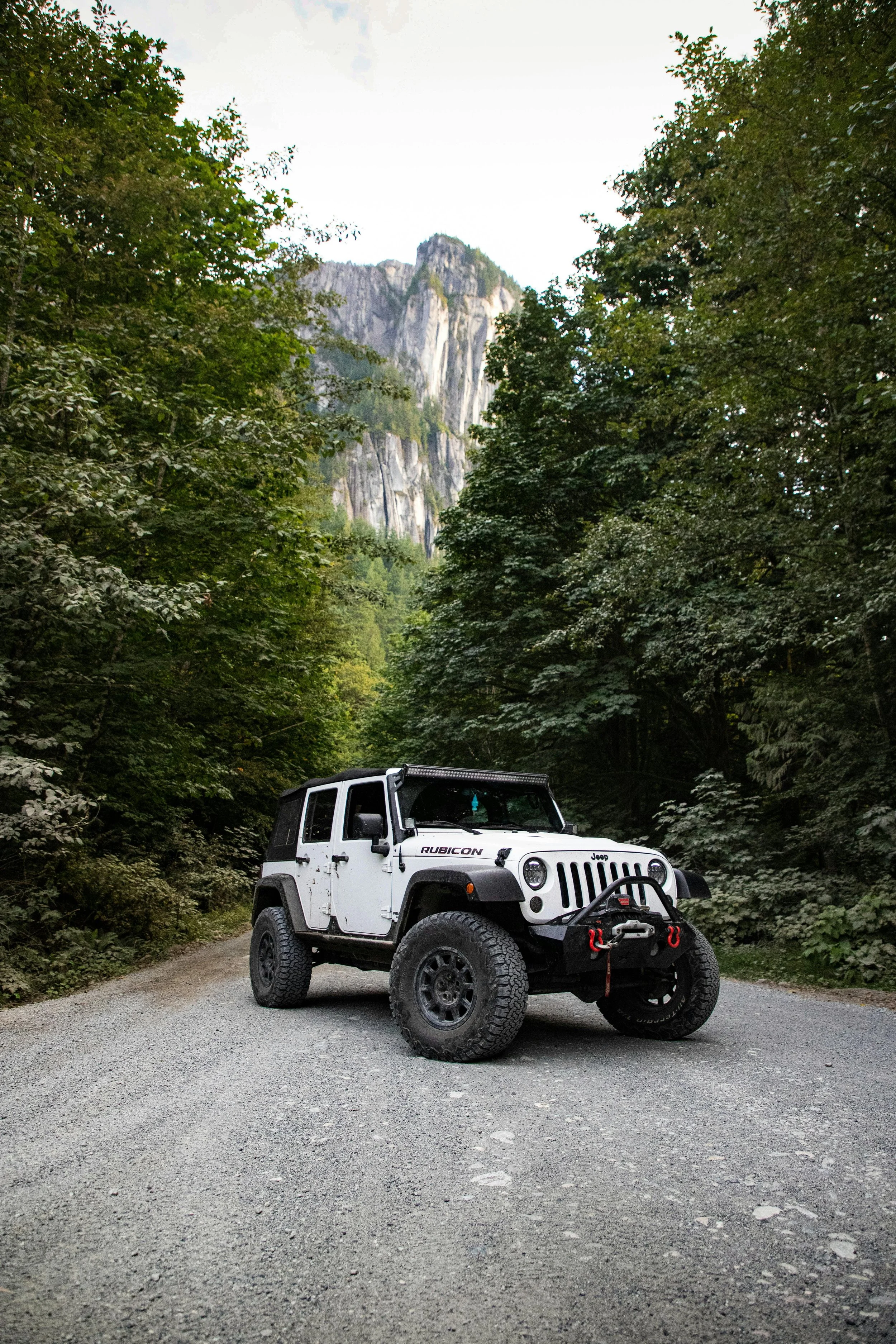 A white Jeep Rubicon parked on a gravel forest road surrounded by lush green trees, with tall rocky mountain cliffs in the background.
