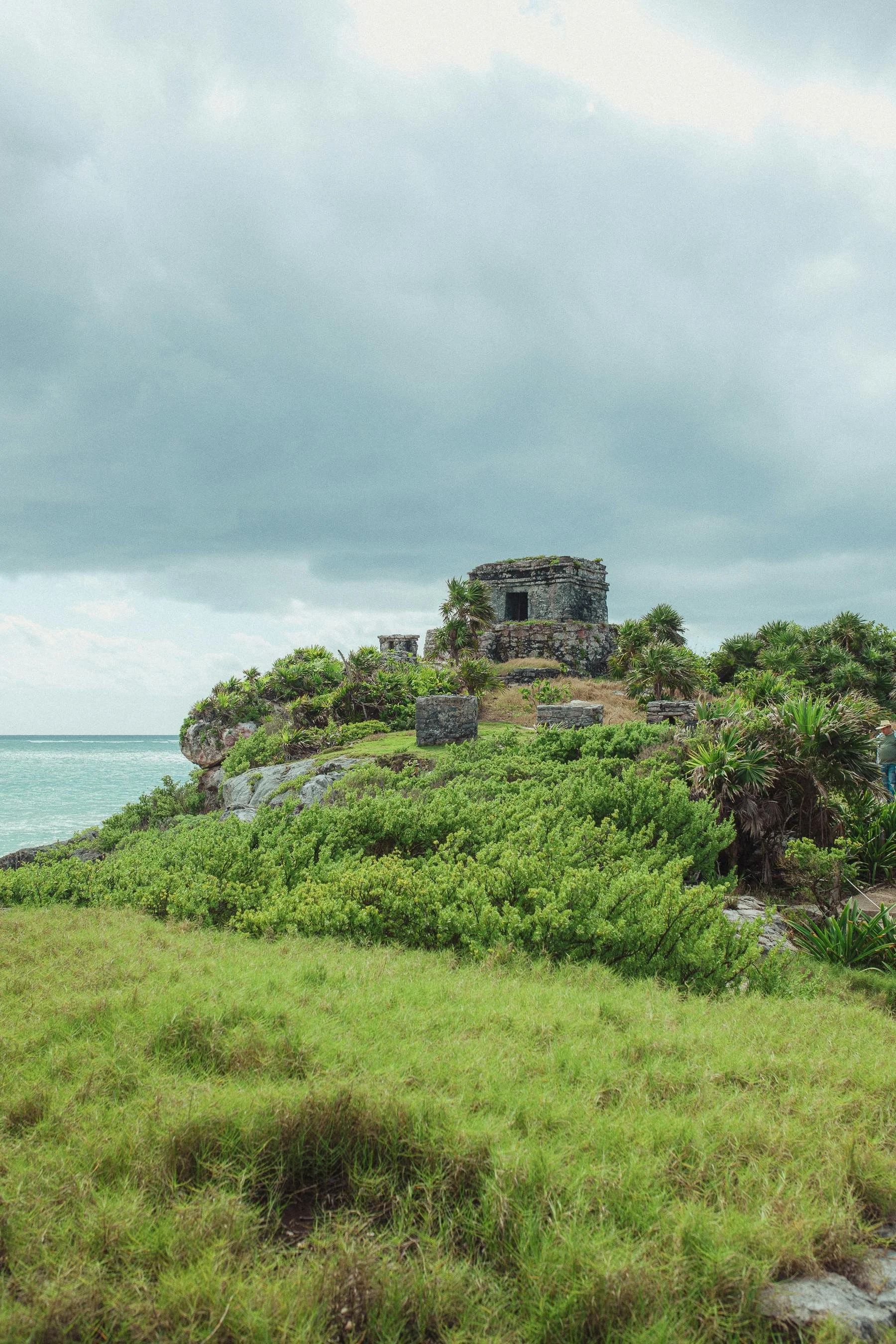 Ancient stone Mayan ruin on a lush green hill near the ocean, under a cloudy sky.