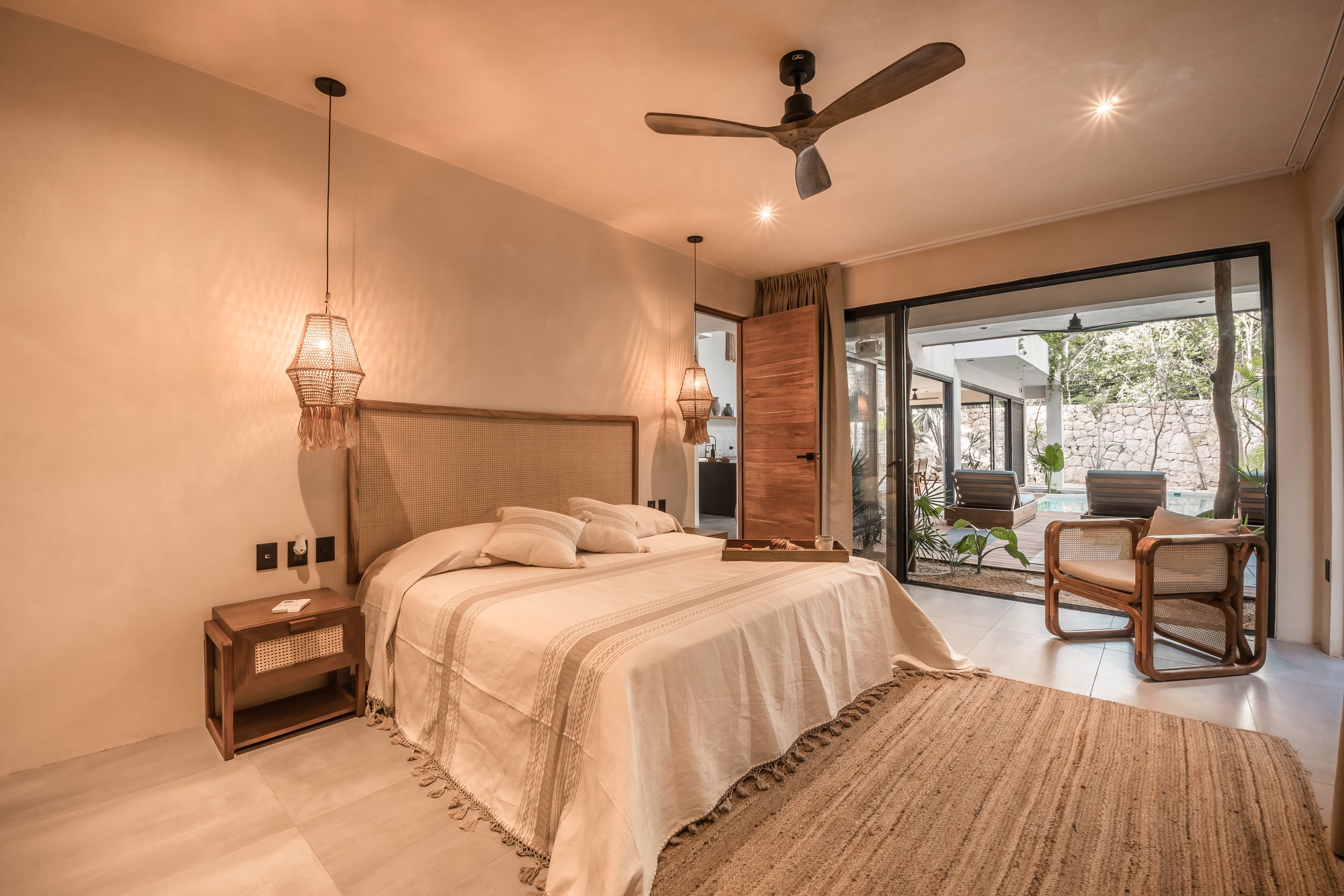 A cozy bedroom with a large bed, hanging pendant lamps, and a ceiling fan. The room has a sliding glass door leading to an outdoor patio with seating and a small pool.