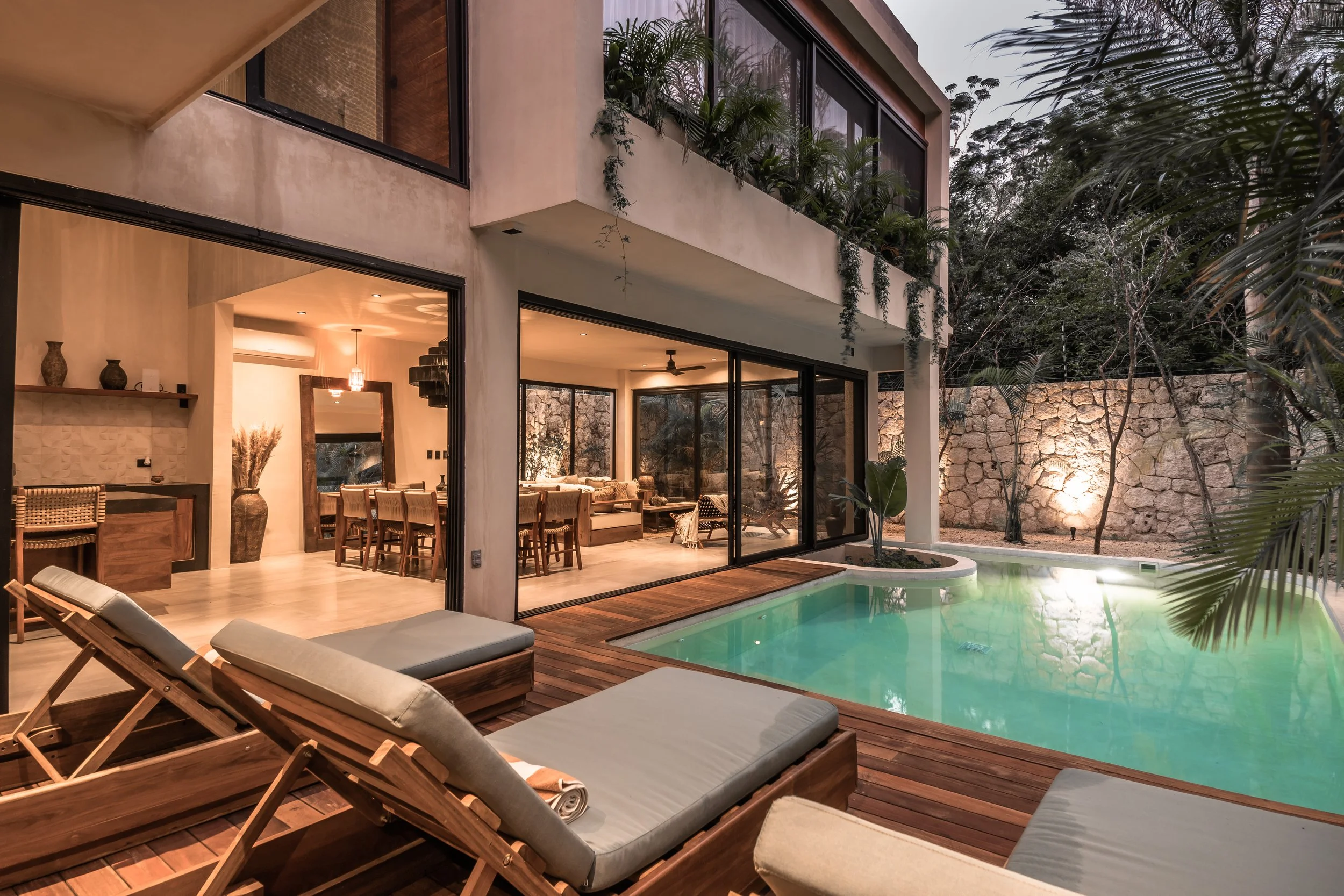 Modern house with large glass windows and a swimming pool with wooden deck and lounge chairs, nighttime setting.