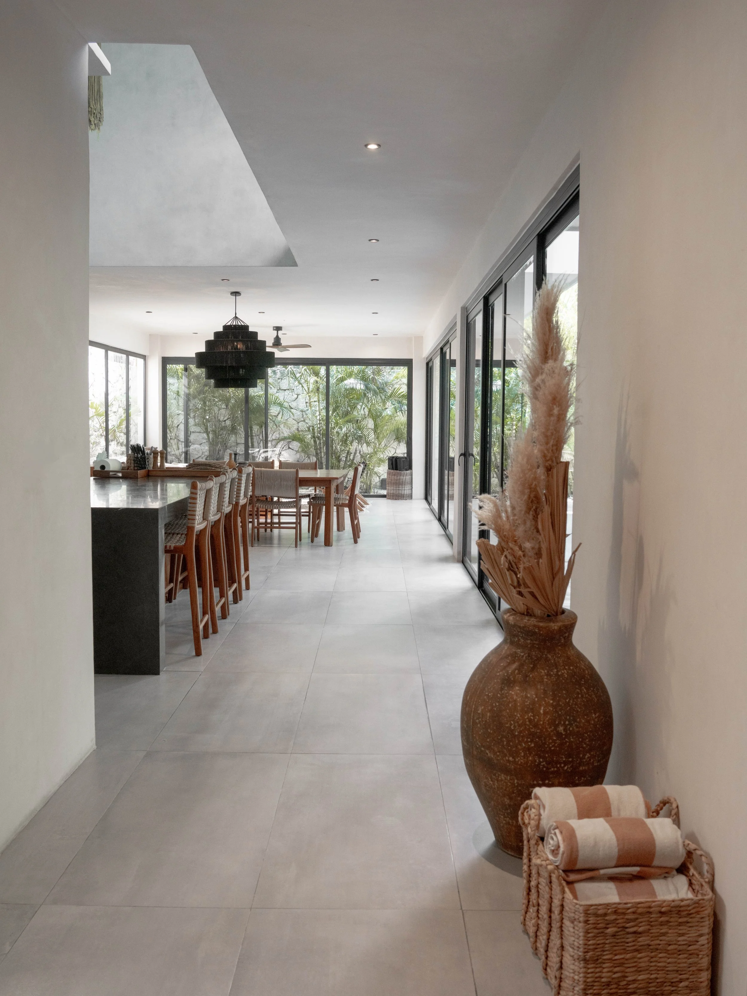 Interior view of a modern dining area with large sliding glass doors, wooden dining table with chairs, a large decorative vase with dried plants, a basket with rolled towels, and greenery outside.