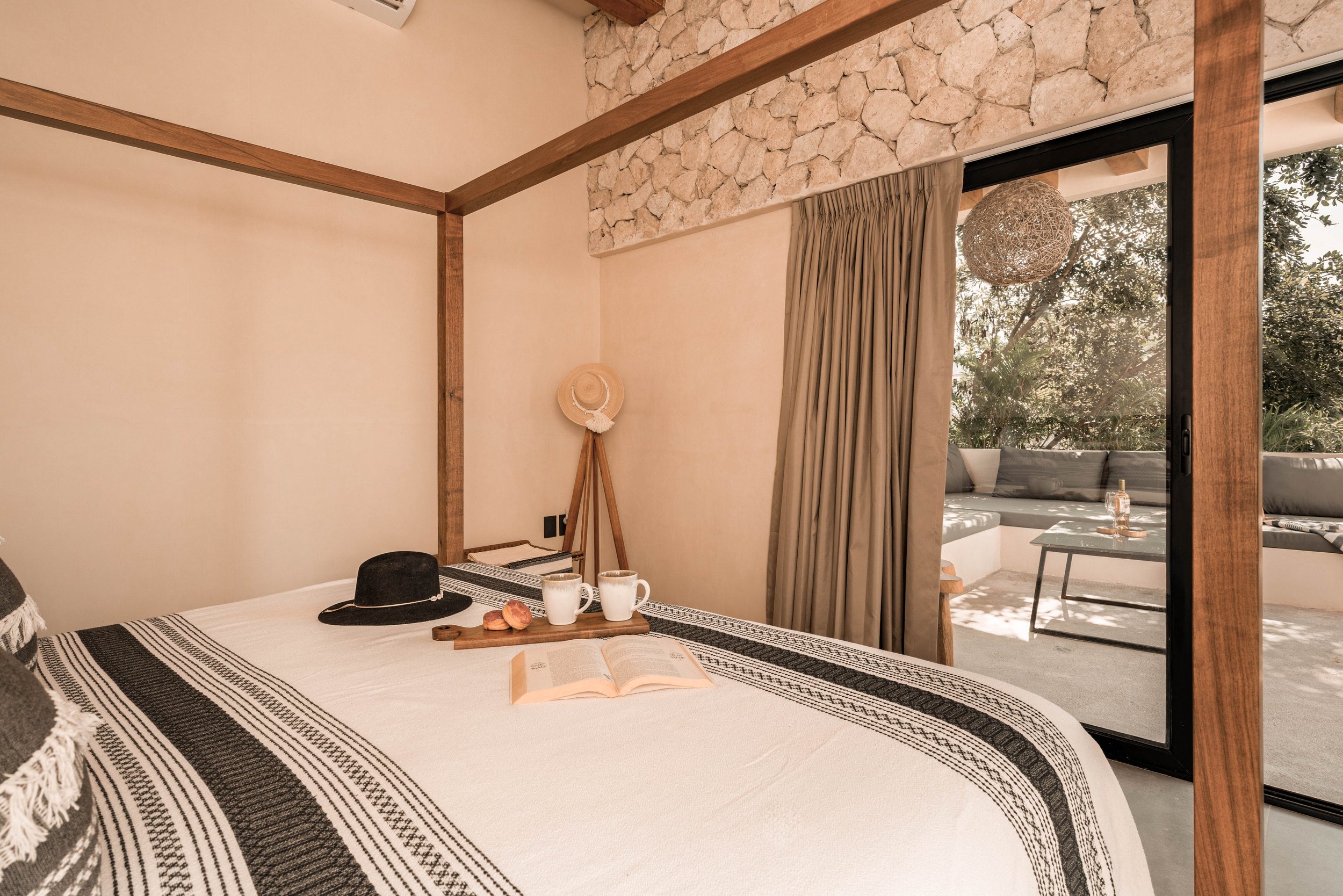 A cozy bedroom with a sliding glass door leading to an outdoor seating area. The bed has a white bedspread with black decorative stripes, and is accessorized with a black wide-brimmed hat, a book, two white mugs, and some pastries on a tray. Nearby, a hat is hung on a wooden tripod stand. The room features beige walls, wood beams, and a stone accent wall above the bed.