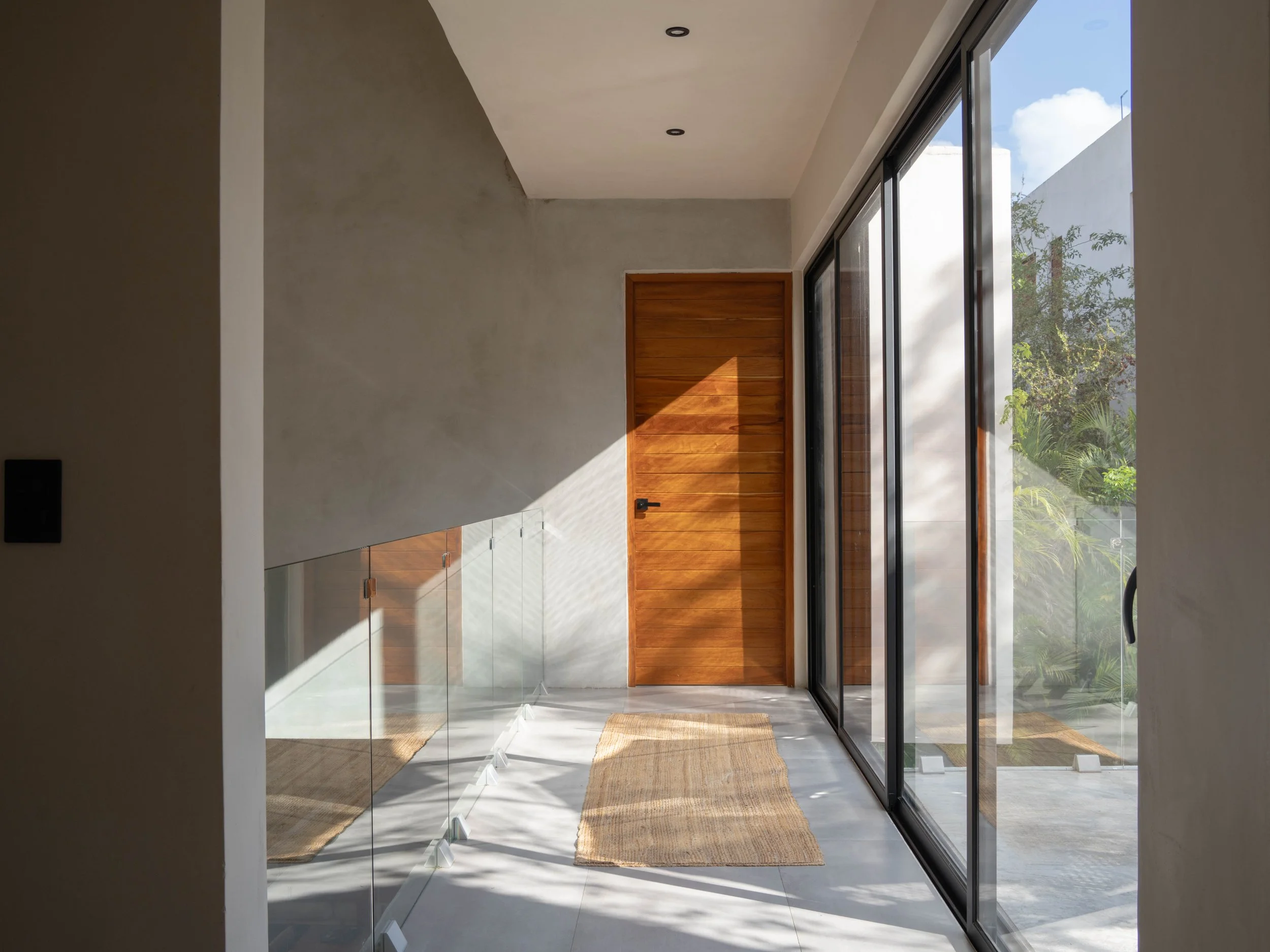 Modern entryway with a wooden door, glass sliding doors, and sunlight creating shadows on the wall and floor.