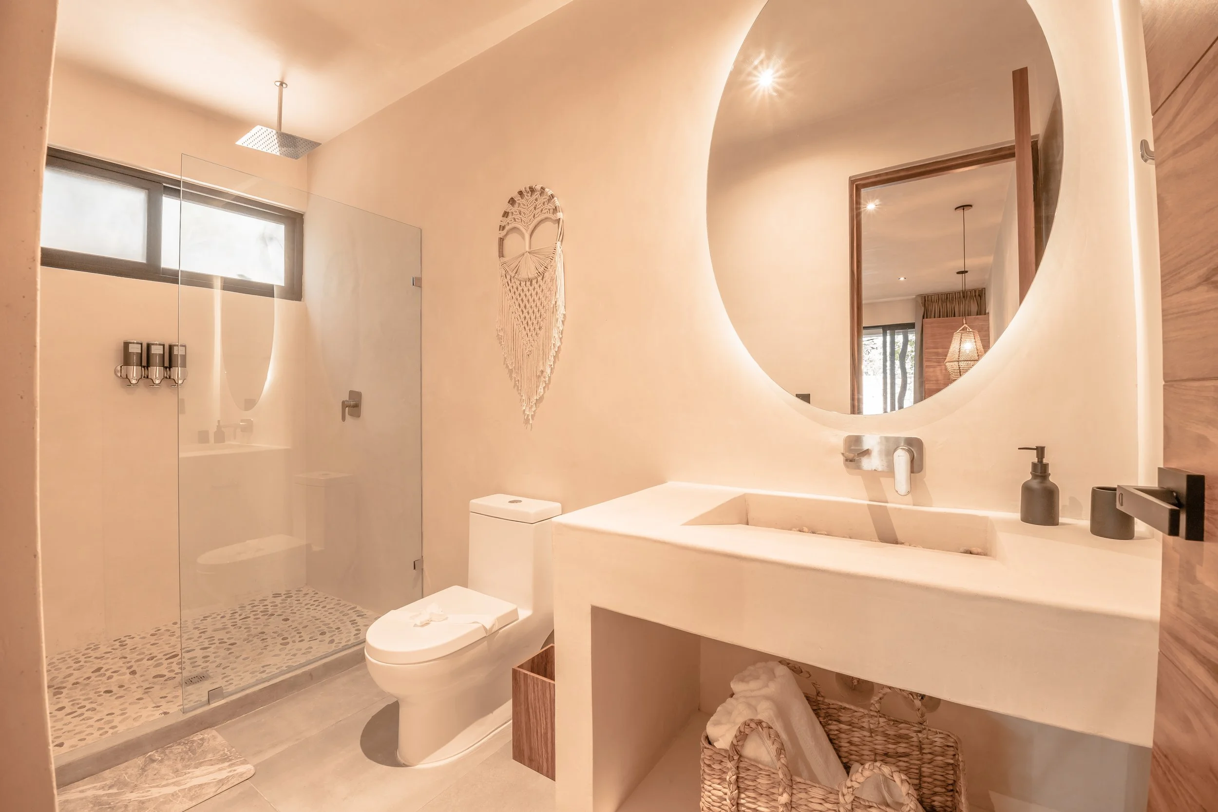 Modern bathroom with round mirror above a concrete sink, a toilet, a glass-enclosed shower, and decorative wall hanging, in neutral tones.