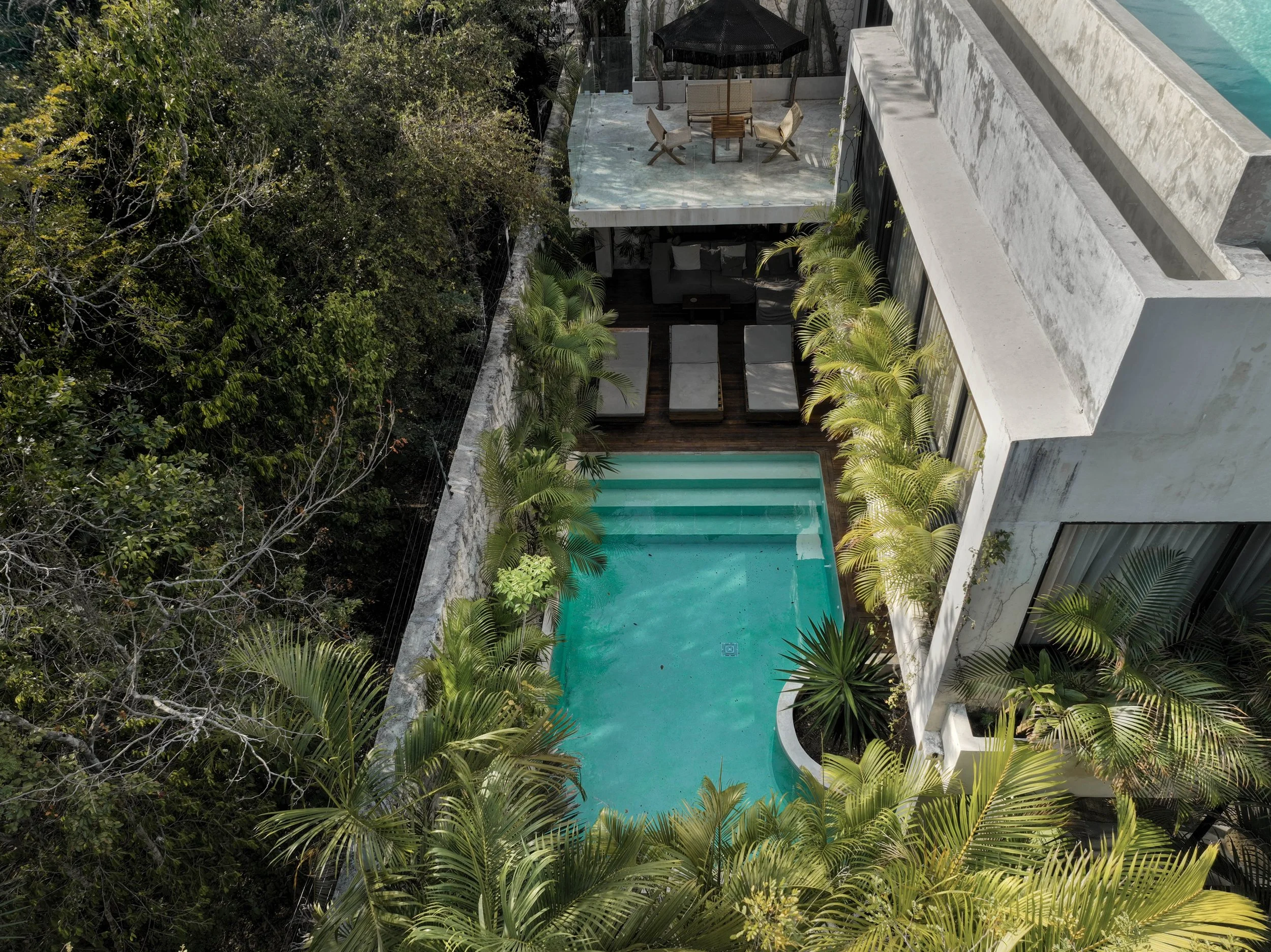Aerial view of a modern building with a private pool surrounded by lush tropical plants and trees, outdoor seating area with tables and chairs under a black umbrella.
