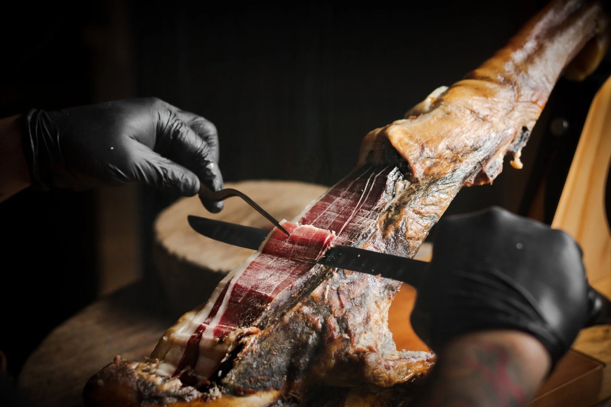 Person in black gloves slices cured ham from a ham leg using a long, sharp knife.