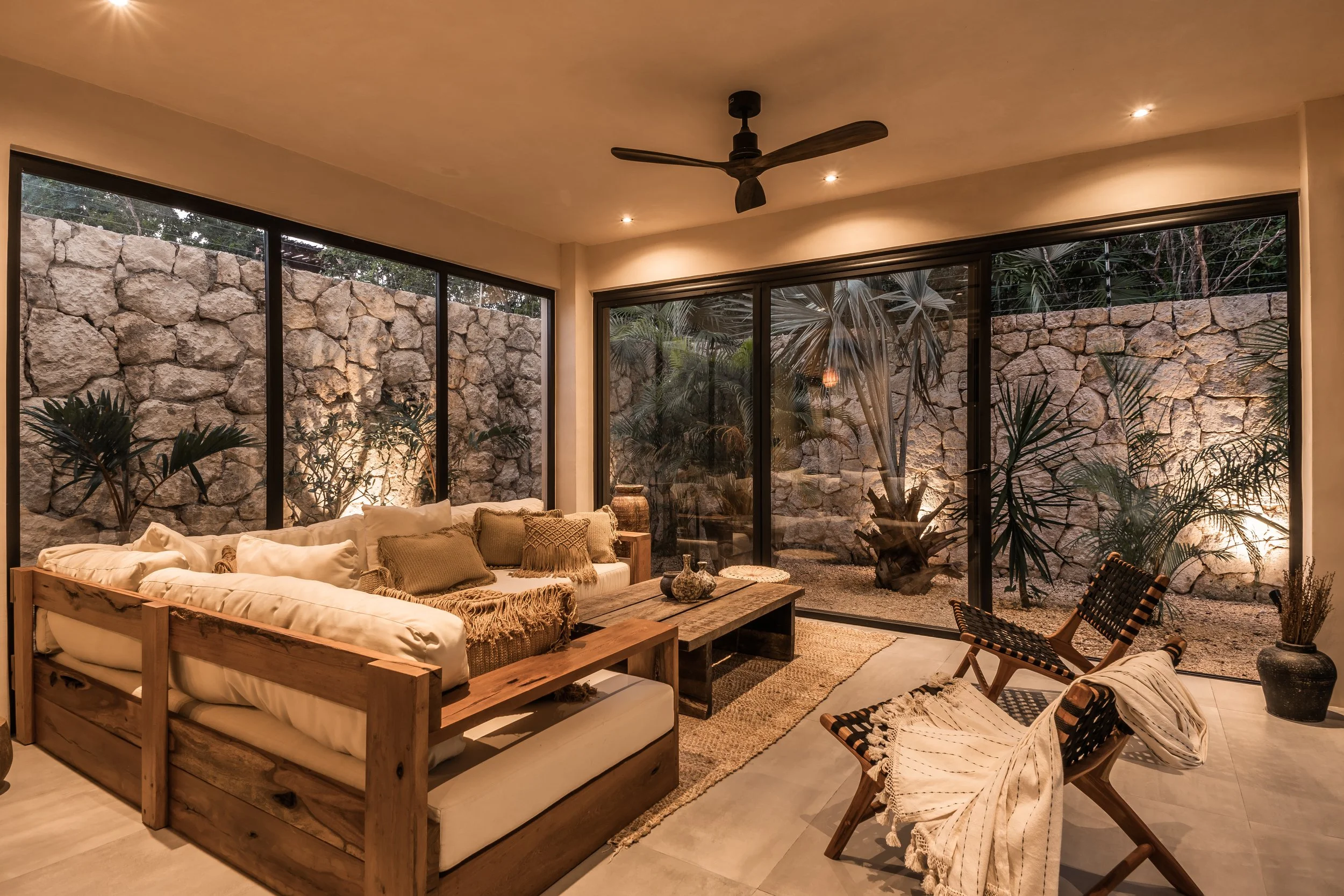 Living room with large glass windows overlooking a stone wall and plants outside, furnished with a white sectional sofa, wooden coffee table, and woven chairs.
