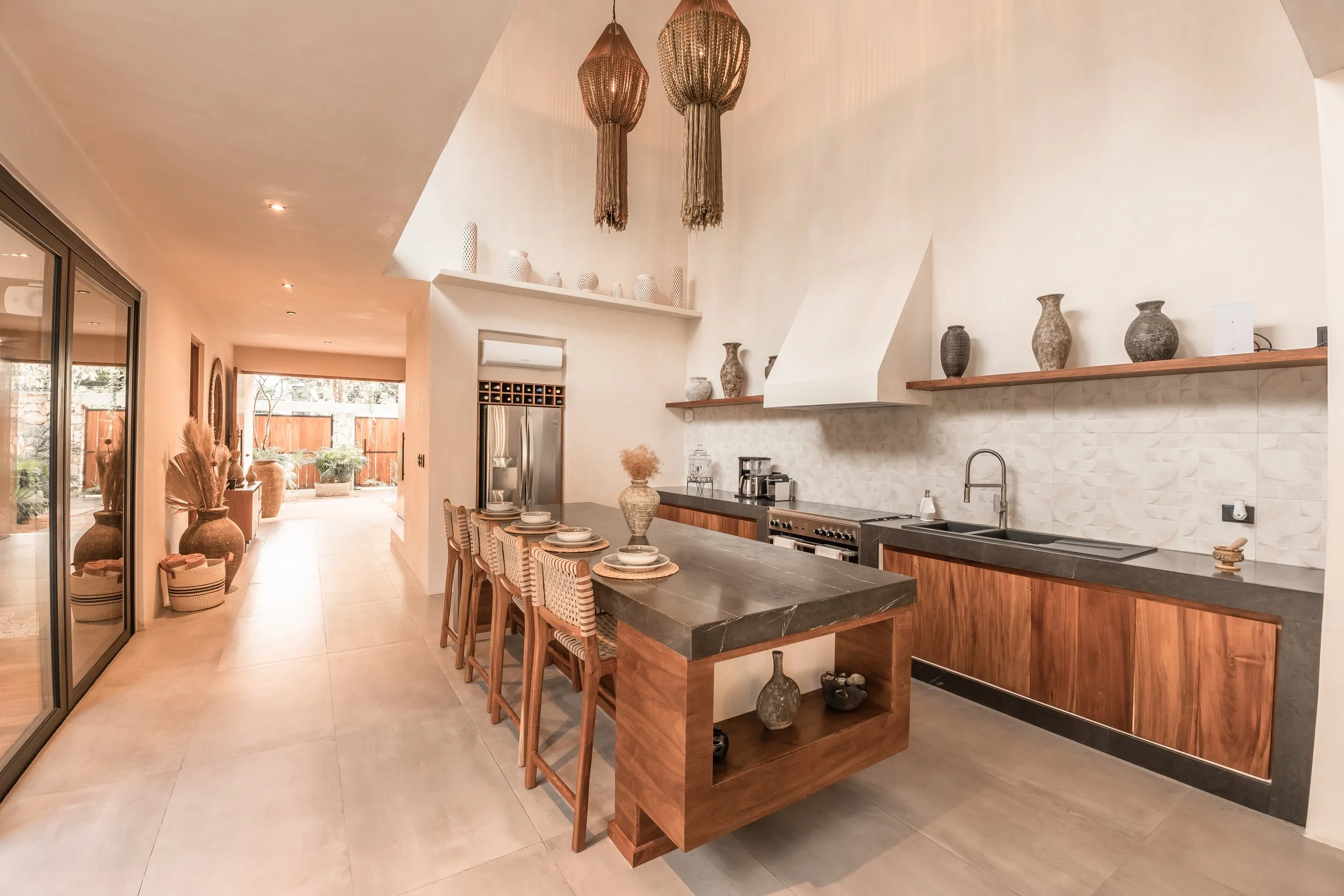 Modern kitchen with wooden cabinetry, black countertop, decorative vases, hanging pendant lights, and an open dining area with bar stools.