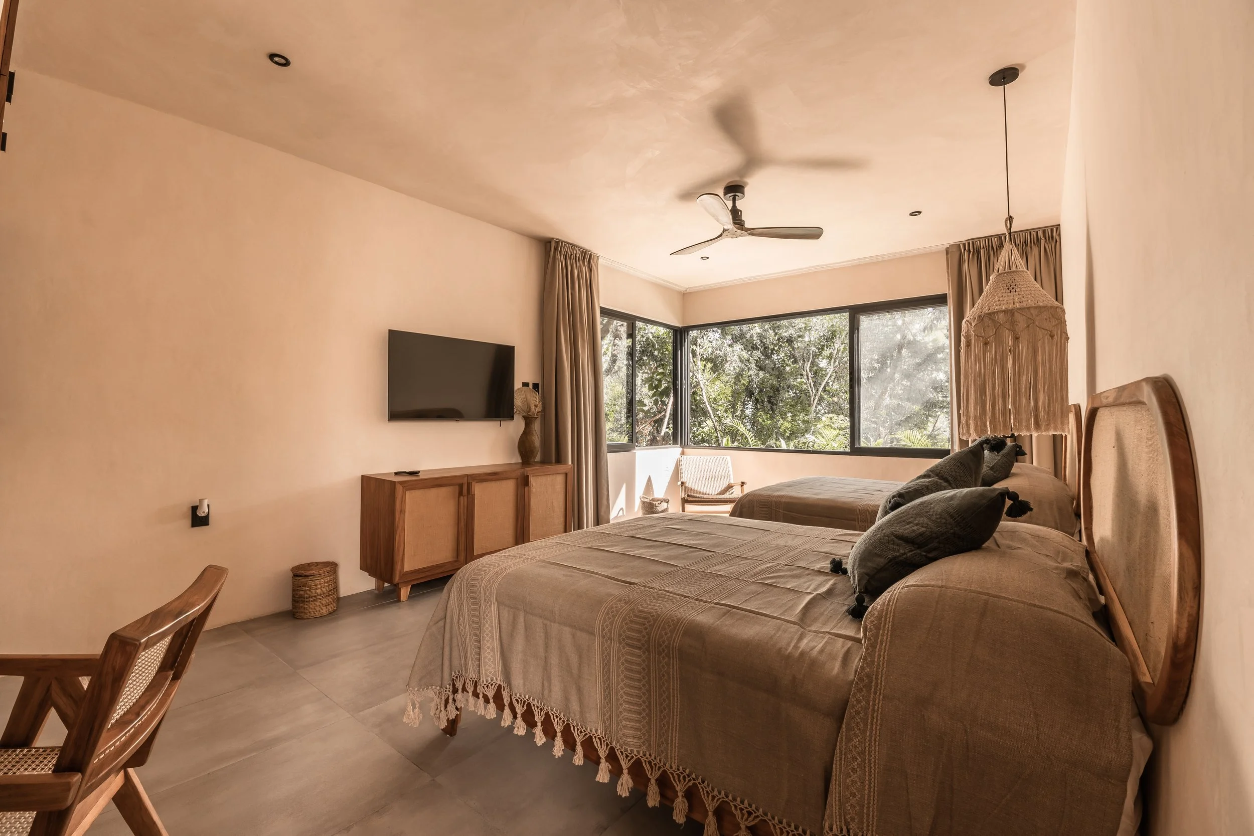 A bedroom with two beds, a wall-mounted flat-screen TV, large windows with curtains, a ceiling fan, and natural scenery outside.