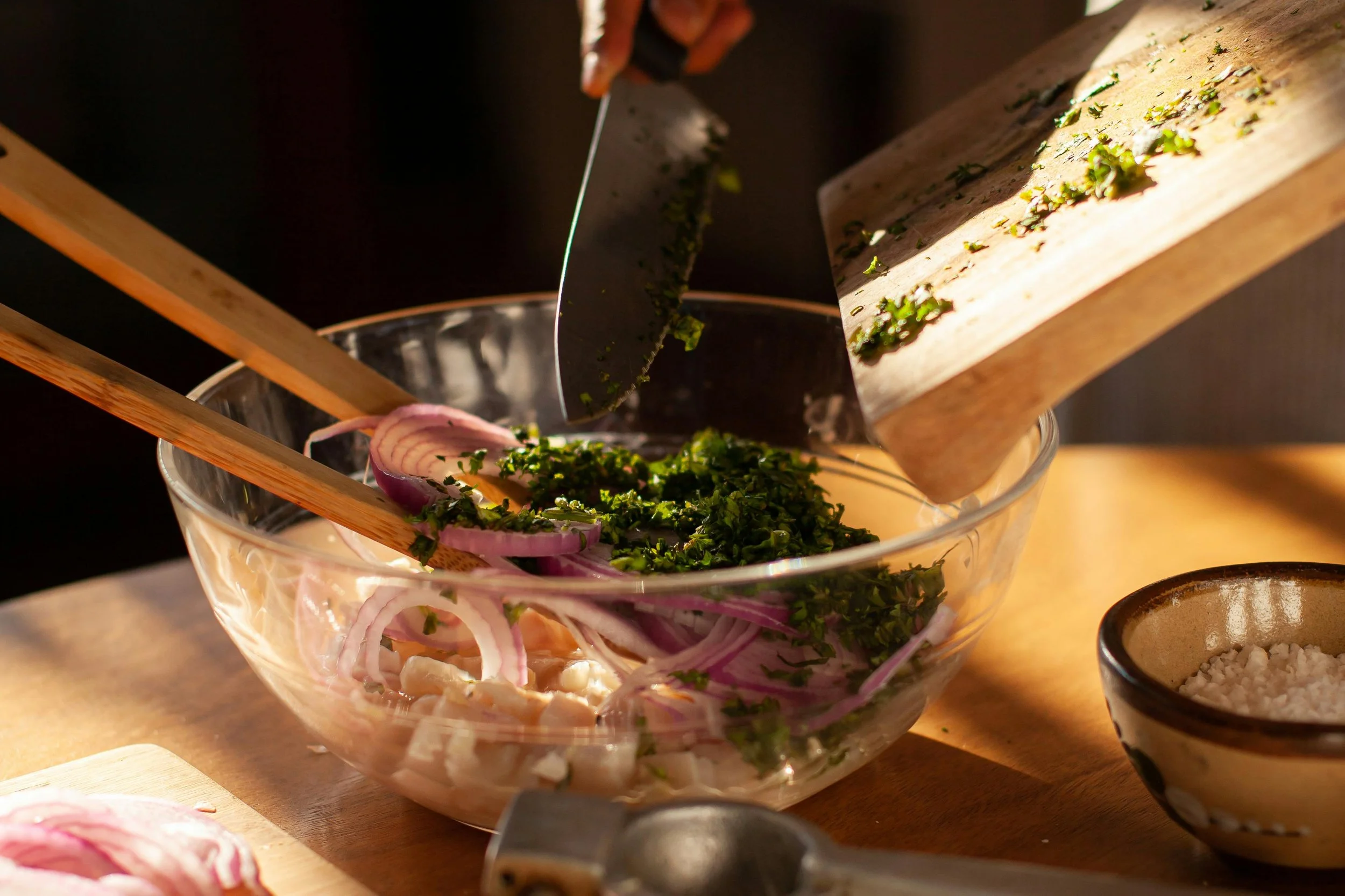 A person chopping fresh herbs over a bowl of chopped onions, with a cutting board and small bowl of salt nearby.