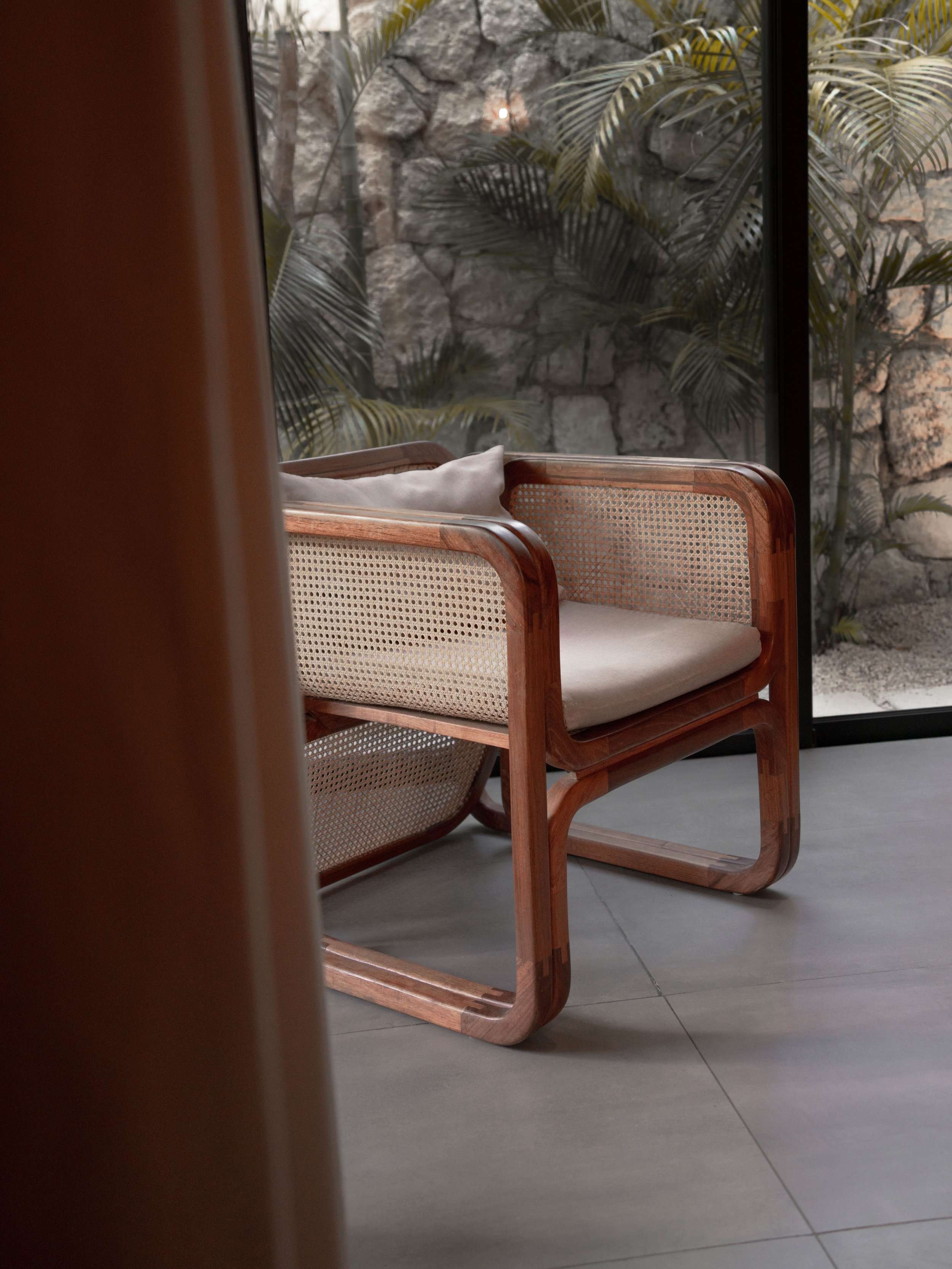 Wooden lounge chair with woven rattan sides and cushion, positioned near a large window with scenic outdoor plants and rocks visible outside.