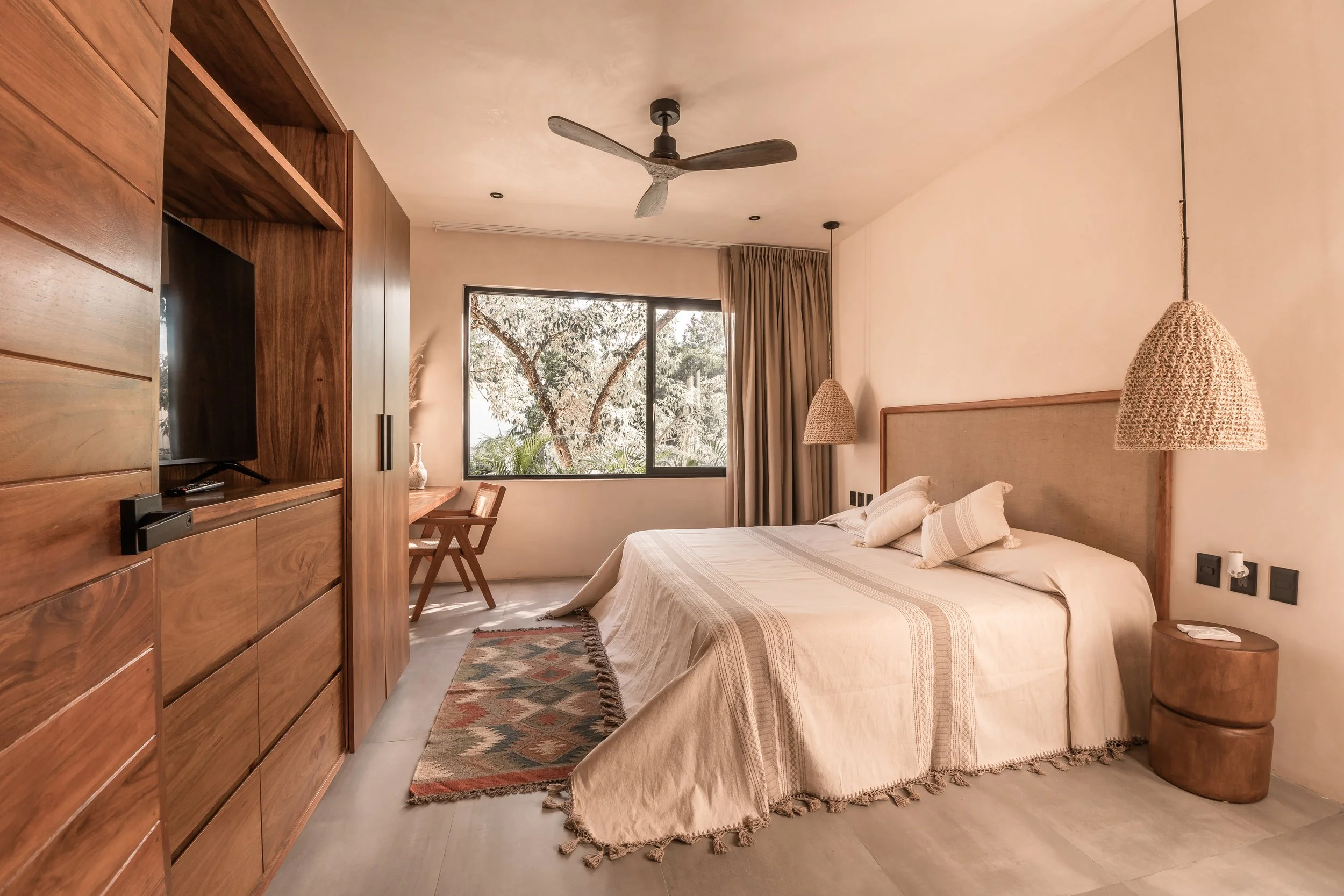 Hotel room with a bed, rattan pendant lights, wooden furniture, a window with trees outside, beige walls, and a small rug.