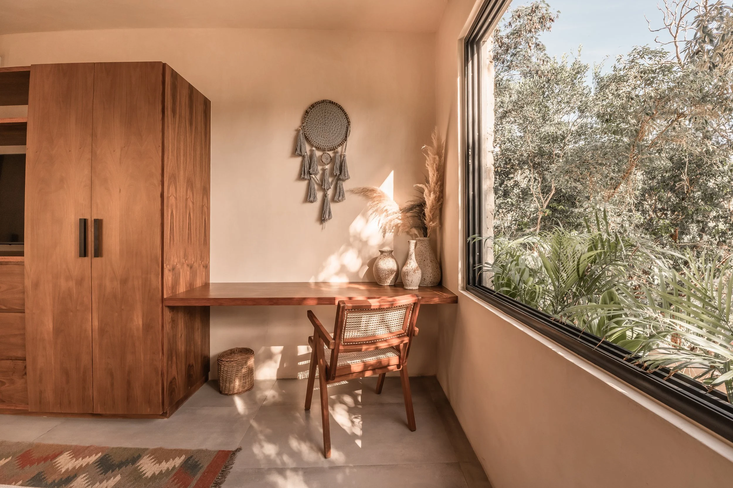 Interior scene with a wooden wardrobe, a small wooden desk with decorative vases, a woven basket underneath, a rattan chair, moon-shaped wall hanging, and a large window revealing greenery outside.
