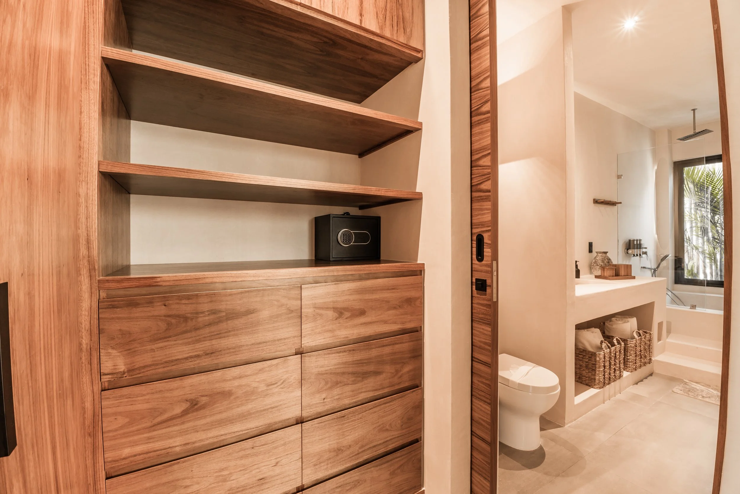 A wooden safe is placed on a wooden dresser within a modern bathroom. The bathroom has a white bathtub, a step, a sink with decor, and baskets underneath, with a window showing outside greenery.