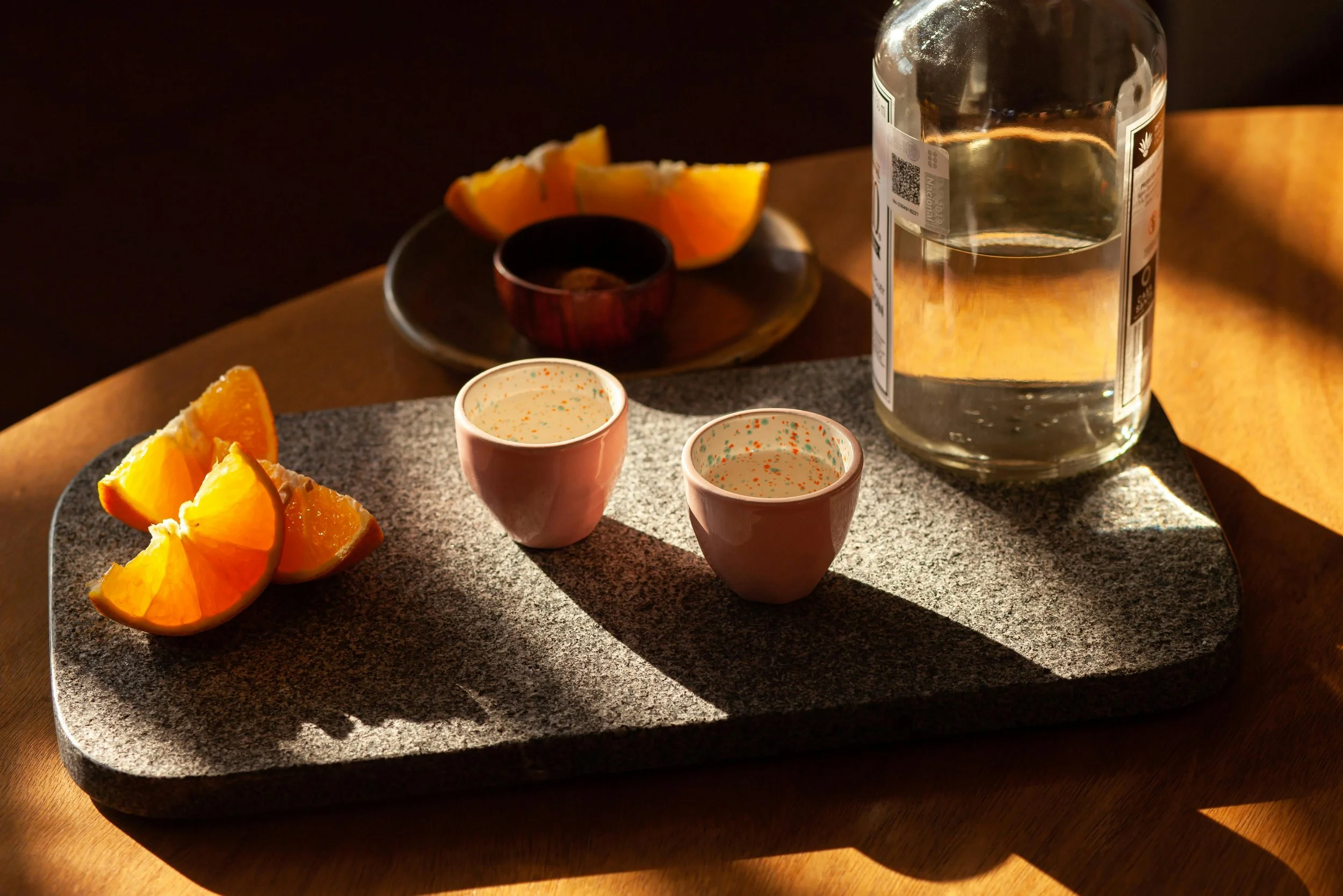 A gray textured serving tray holds orange wedges and two small pink cups, with a large clear bottle of water, and a plate of orange wedges in the background. Sunlight casts shadows on the tray.
