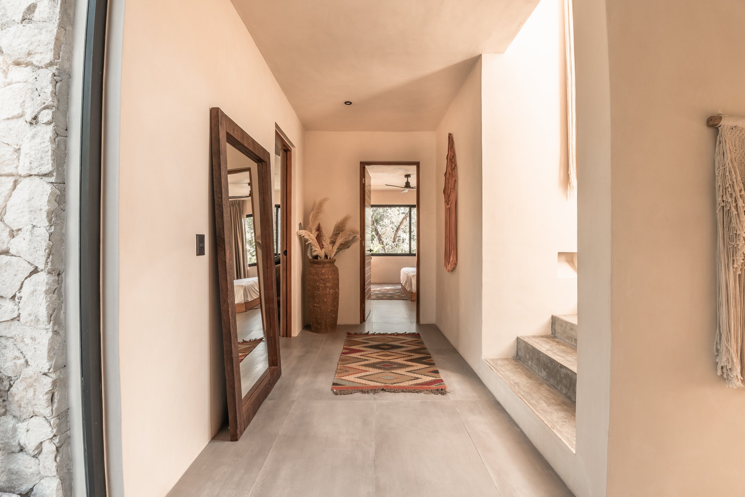 A hallway with cream-colored walls, decorated with a large mirror with a wooden frame, a woven rug, tall floor vase with pampas grass, and wall hanging art; leading into a bright bedroom with large window and ceiling fan.
