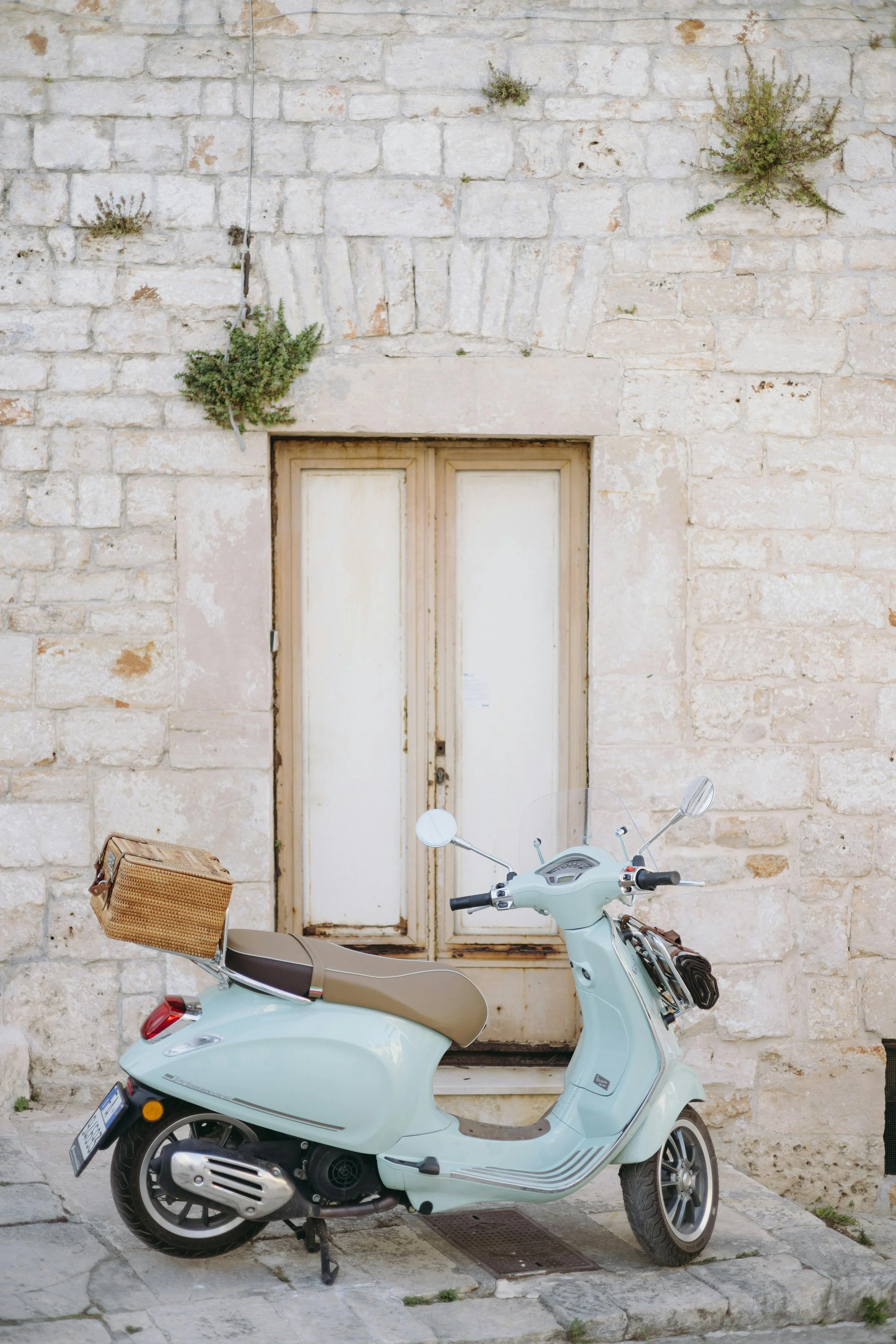 A light blue vintage scooter parked on a cobblestone street in front of a weathered cream-colored stone wall with a closed wooden door and some small green plants growing above.