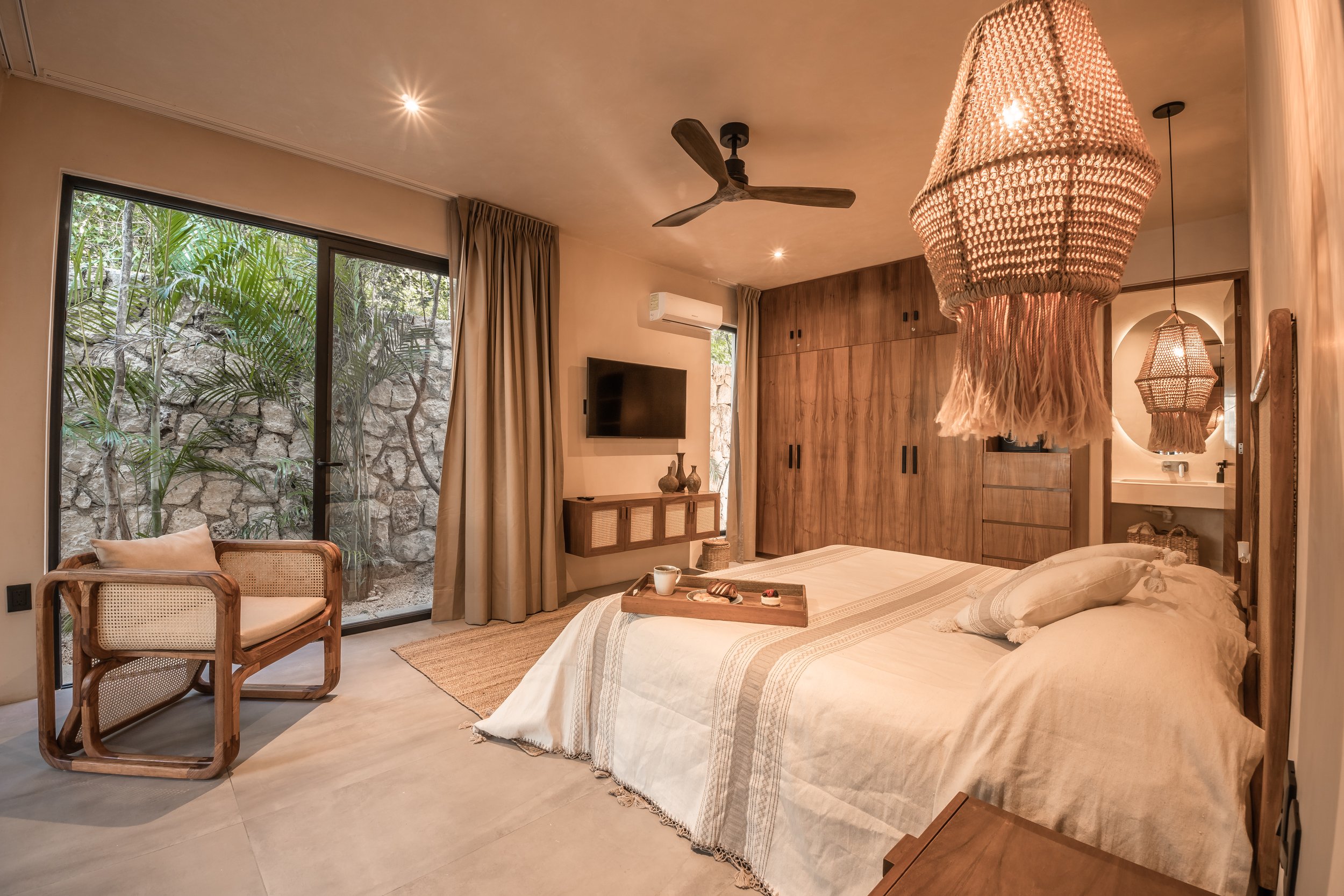 Cozy bedroom with a large bed, wooden wardrobe, wall-mounted TV, armchair, beige curtains, stone wall outside glass door, hanging woven lamps, and natural decor.