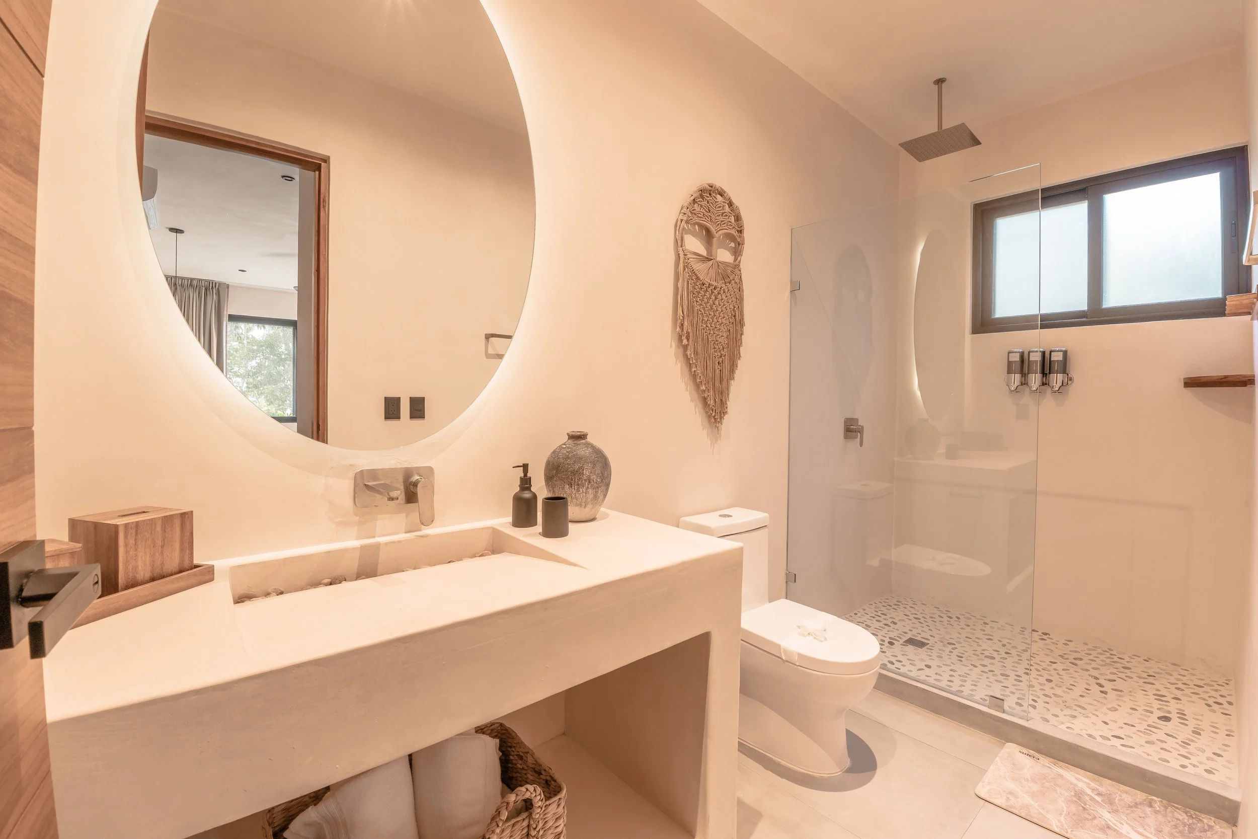 Modern bathroom with a round mirror, beige sink, decorative vases, wall art, toilet, and glass shower with pebble tile floor.