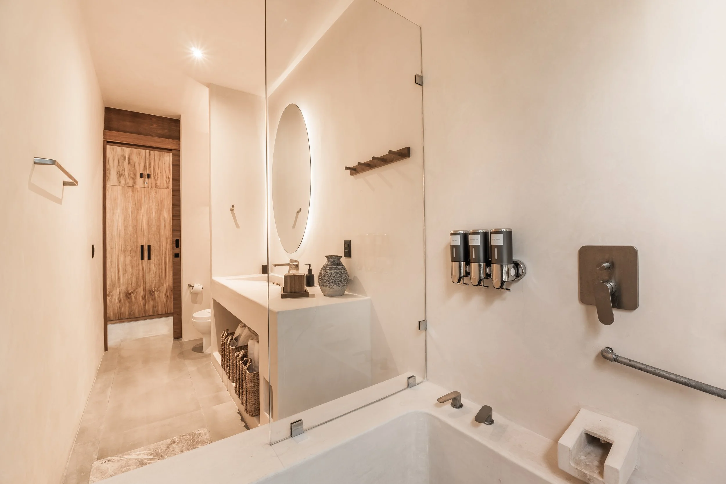 Modern minimalist bathroom with a white bathtub, wall-mounted soap dispensers, a mirror, and wooden accents.