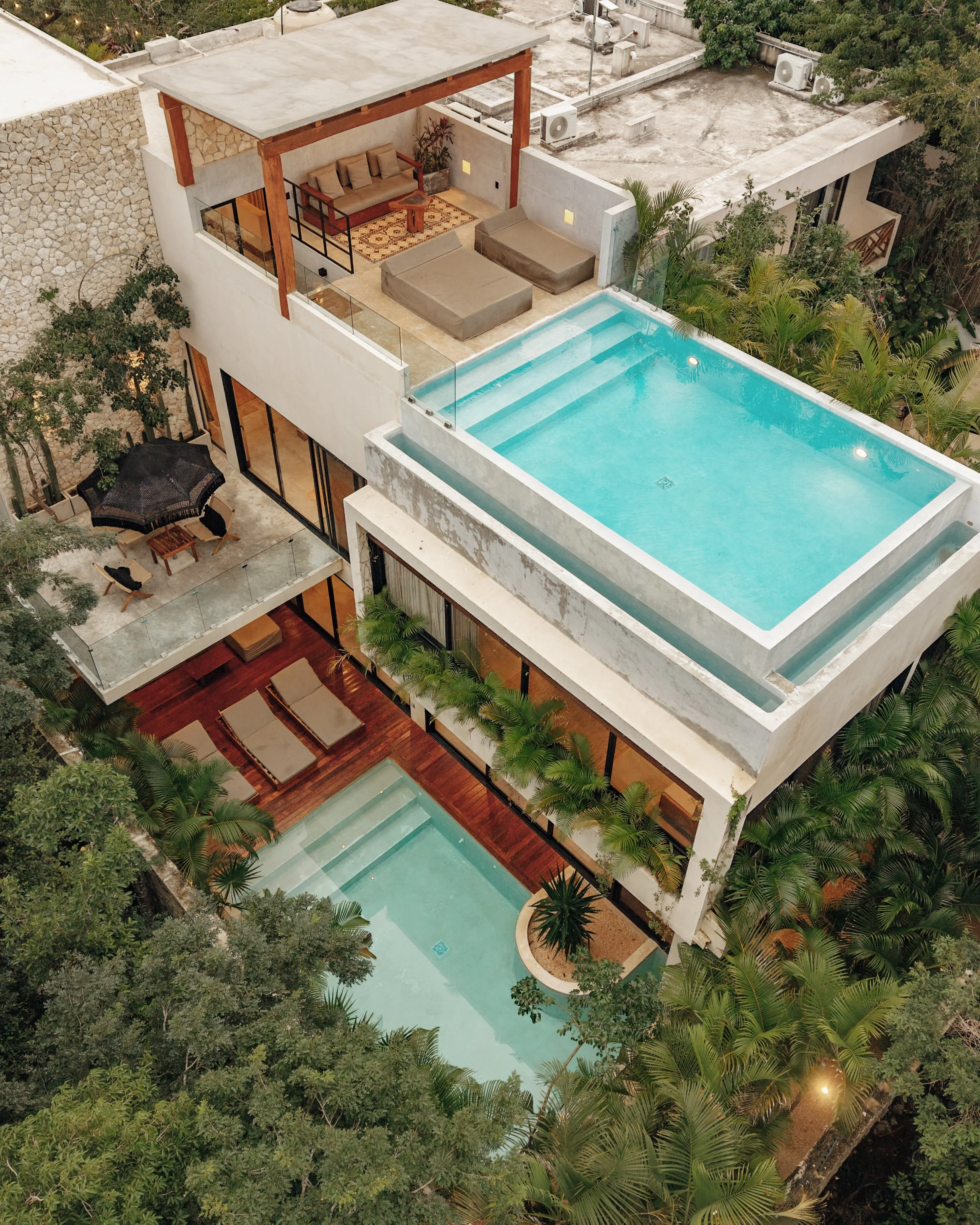 An aerial view of a modern multi-level house with two swimming pools, outdoor seating areas, lush greenery, and various outdoor furniture.