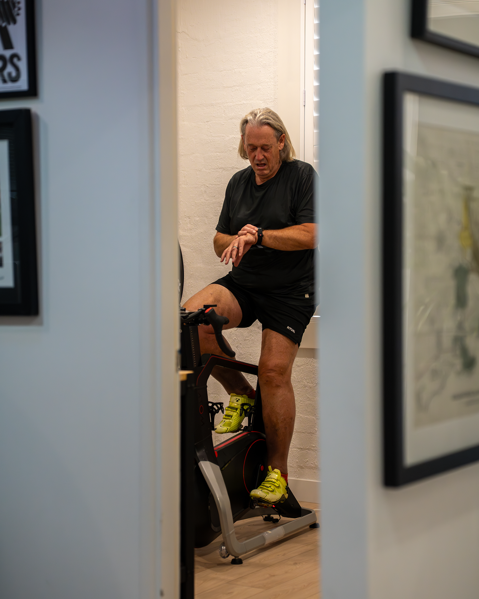 Older man in black athletic wear and yellow running shoes riding a stationary exercise bike indoors.