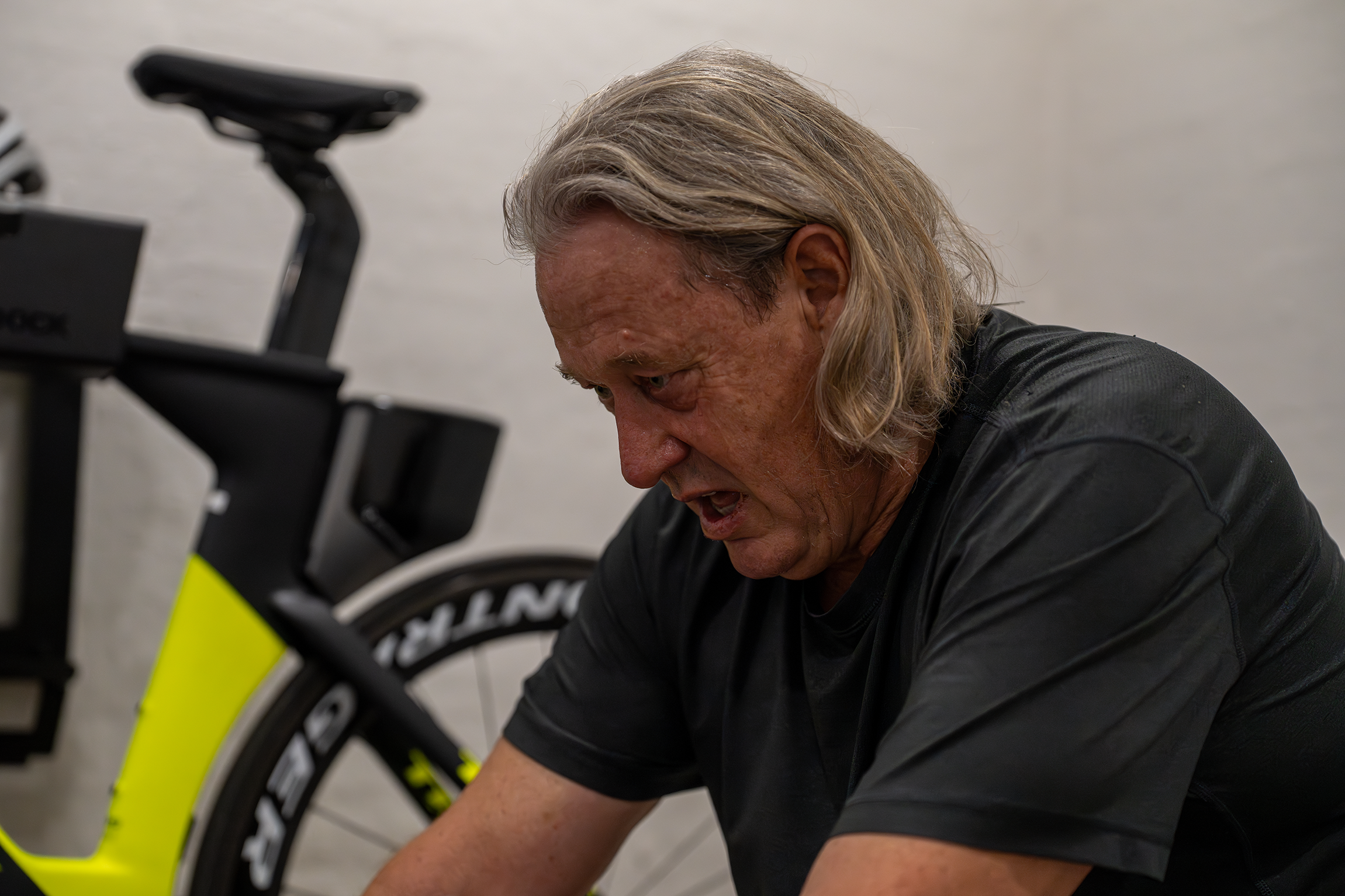 An older man with long gray hair and a Black T-shirt struggling during a workout next to a yellow and black bicycle.