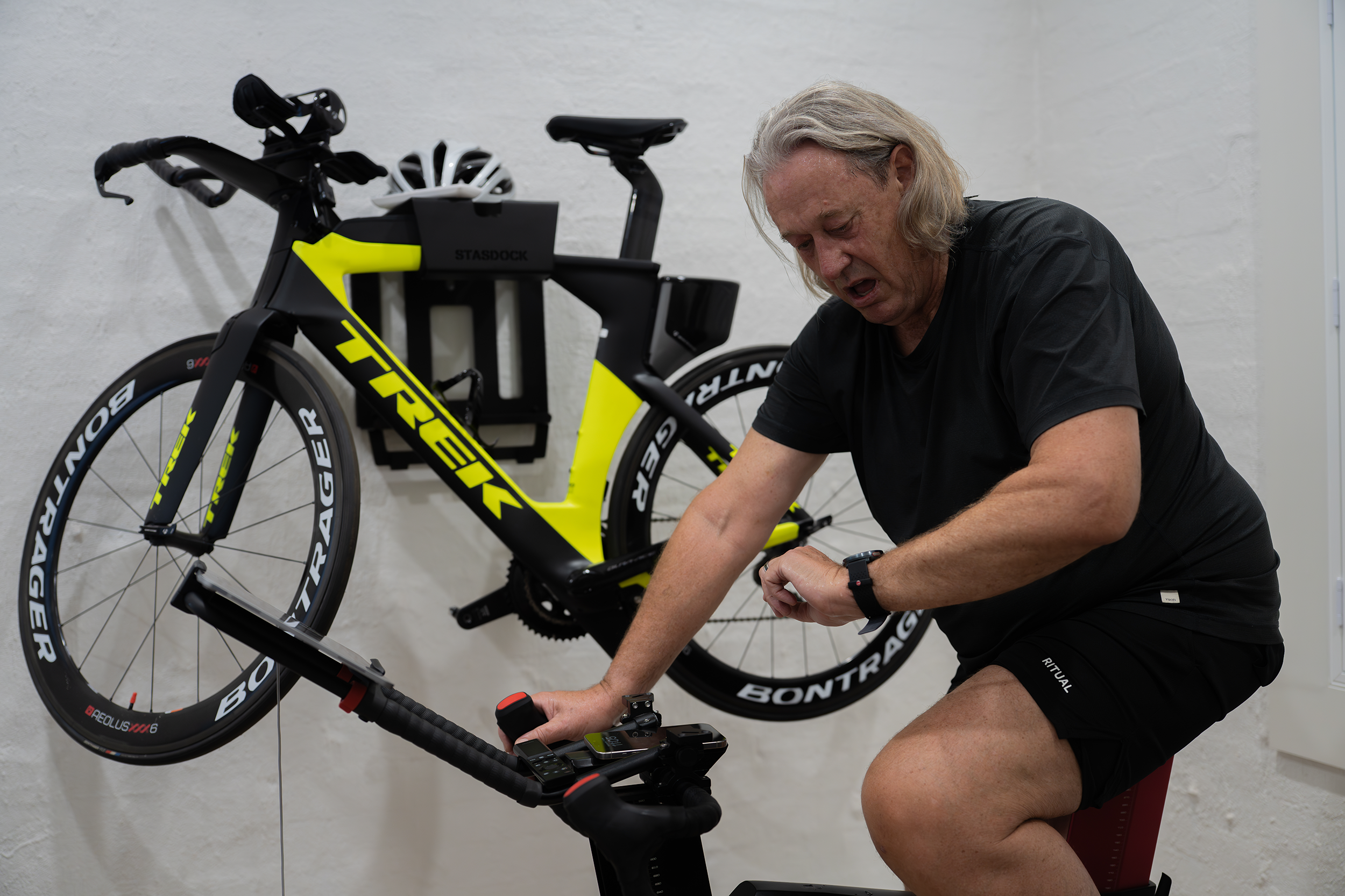 A man with gray hair in black athletic clothing, sitting on a stationary bike, appears exhausted or strained, with a yellow and black Trek racing bicycle mounted on the wall behind him.