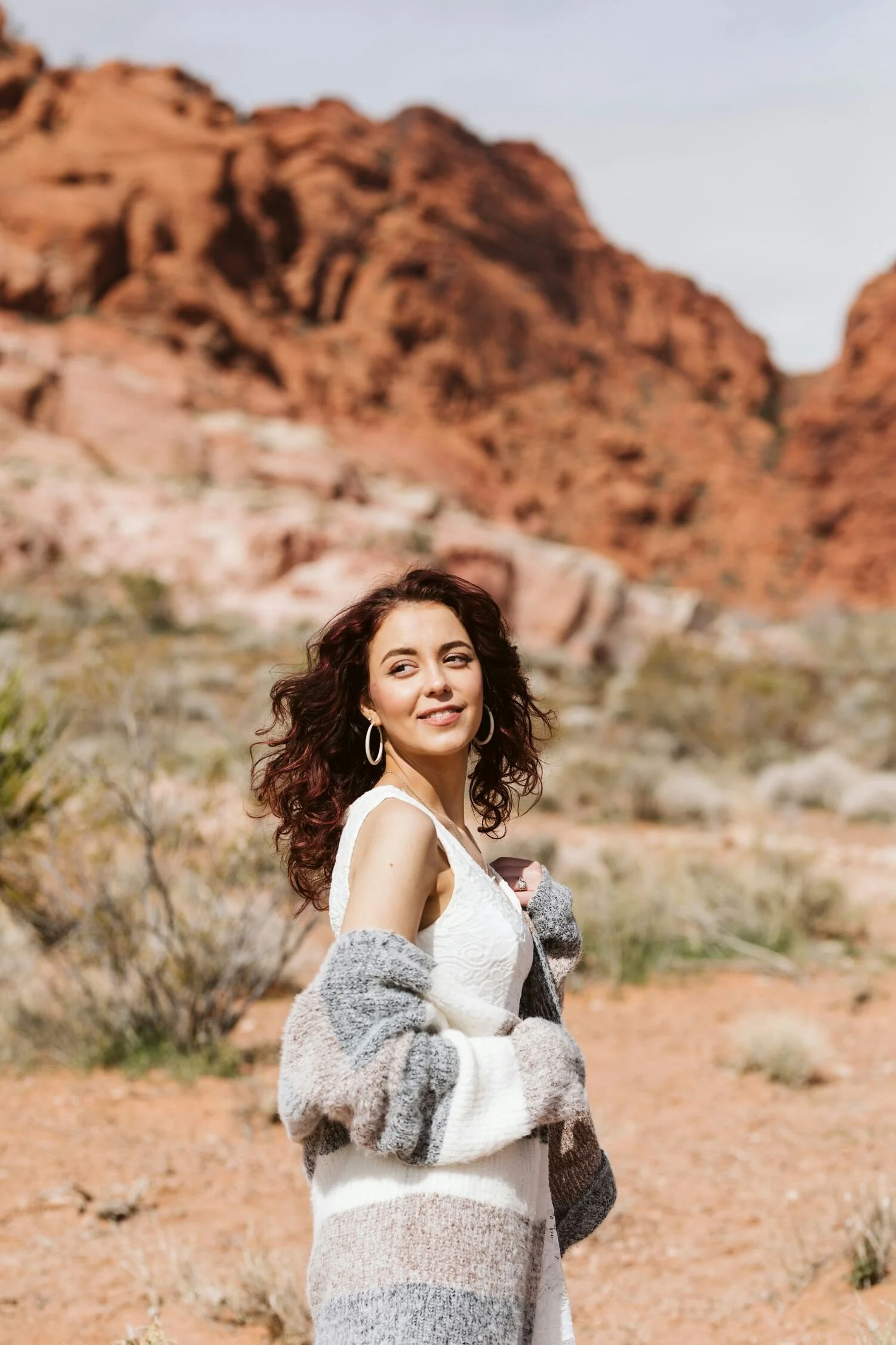 Young woman with dark, curly hair wearing a white dress and a multicolored knit cardigan, standing in a desert landscape with red rock formations in the background.